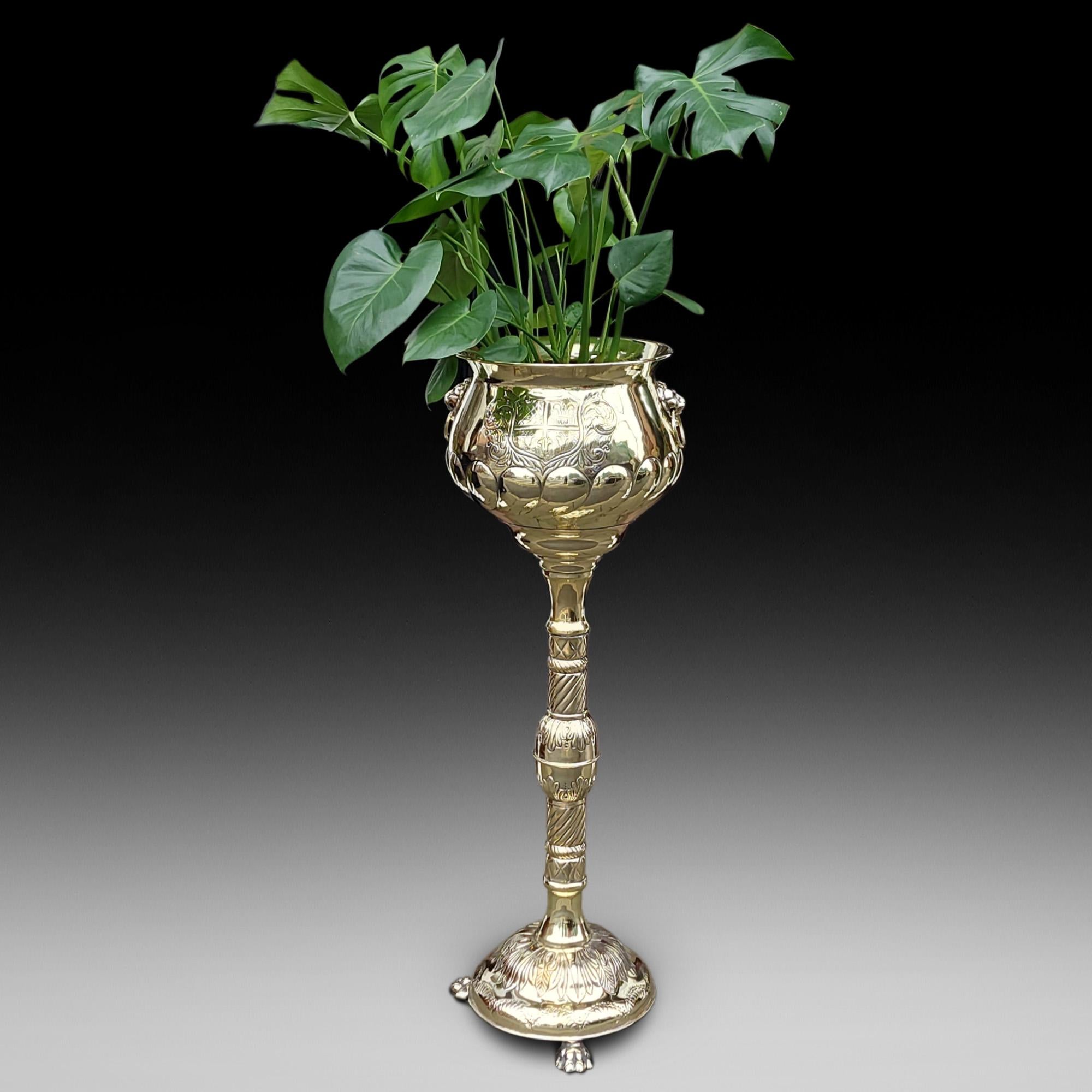 High Victorian Brass Pedestal Jardinière Planter, having relief decoration depicting an armorial crest and lion mask handles to either side, on an ornately tooled column, the plinth base with lion’s paw feet - 13.5
