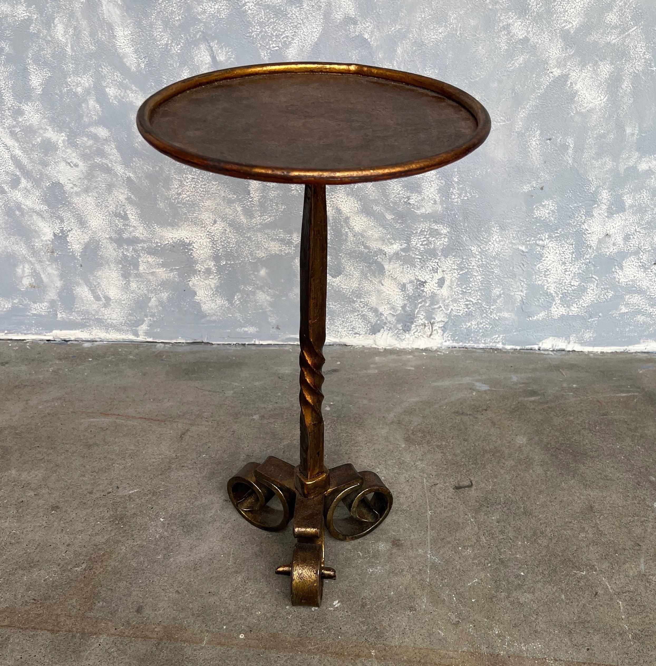 This small intricately decorated gilt iron drinks table features a tripod base complete with curled feet. The central stem presents an intriguing twist detail, supporting a gilt round top encased in a simple rolled ringed border. A hand-applied gold