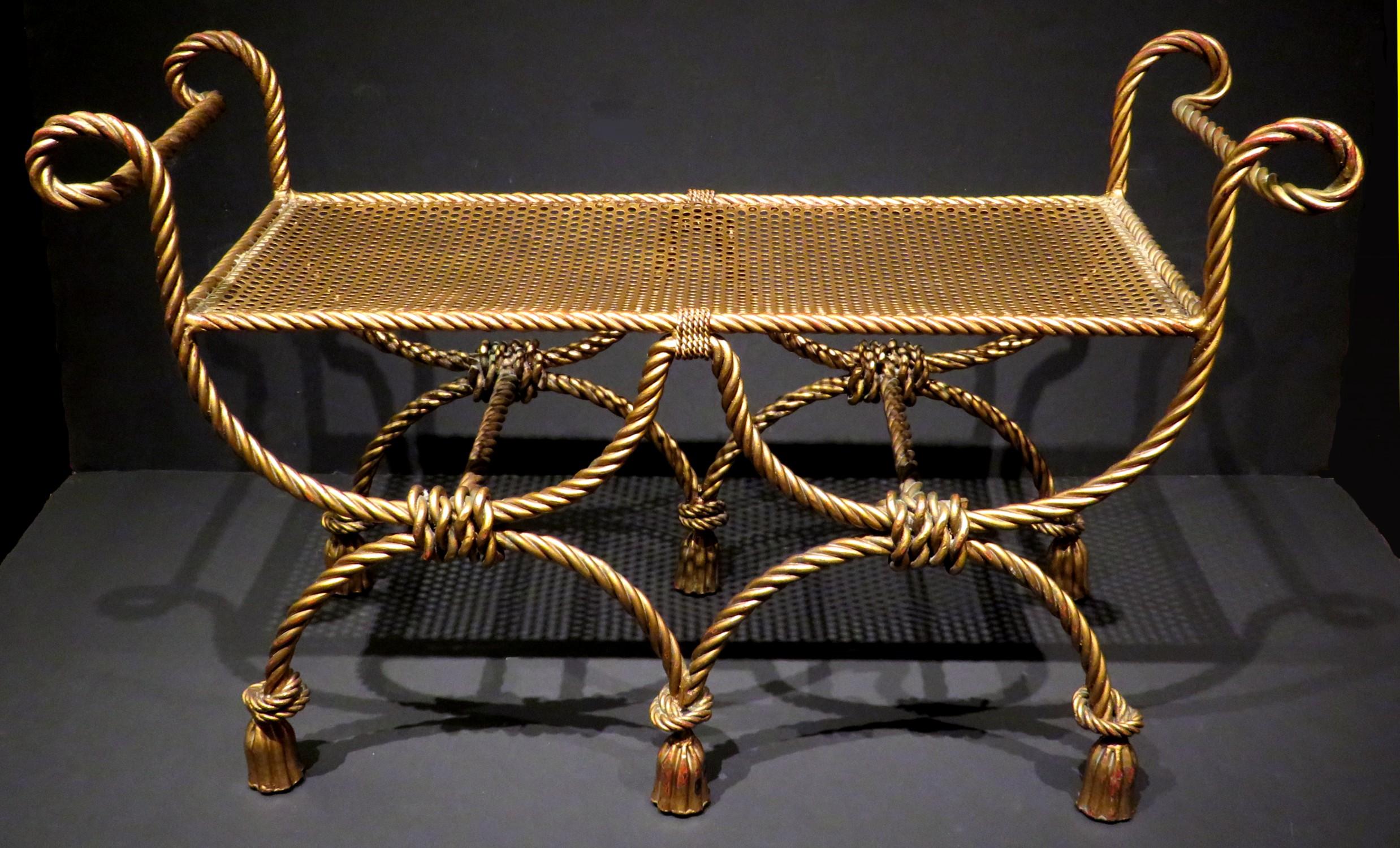 A very attractive & highly stylized gilt-patinated wrought iron bench, the seat raised upon stylized double curule shaped supports in the shape of faux rope & tasseled swags, terminating to six tassel shaped feet. 
A charming accent piece that's