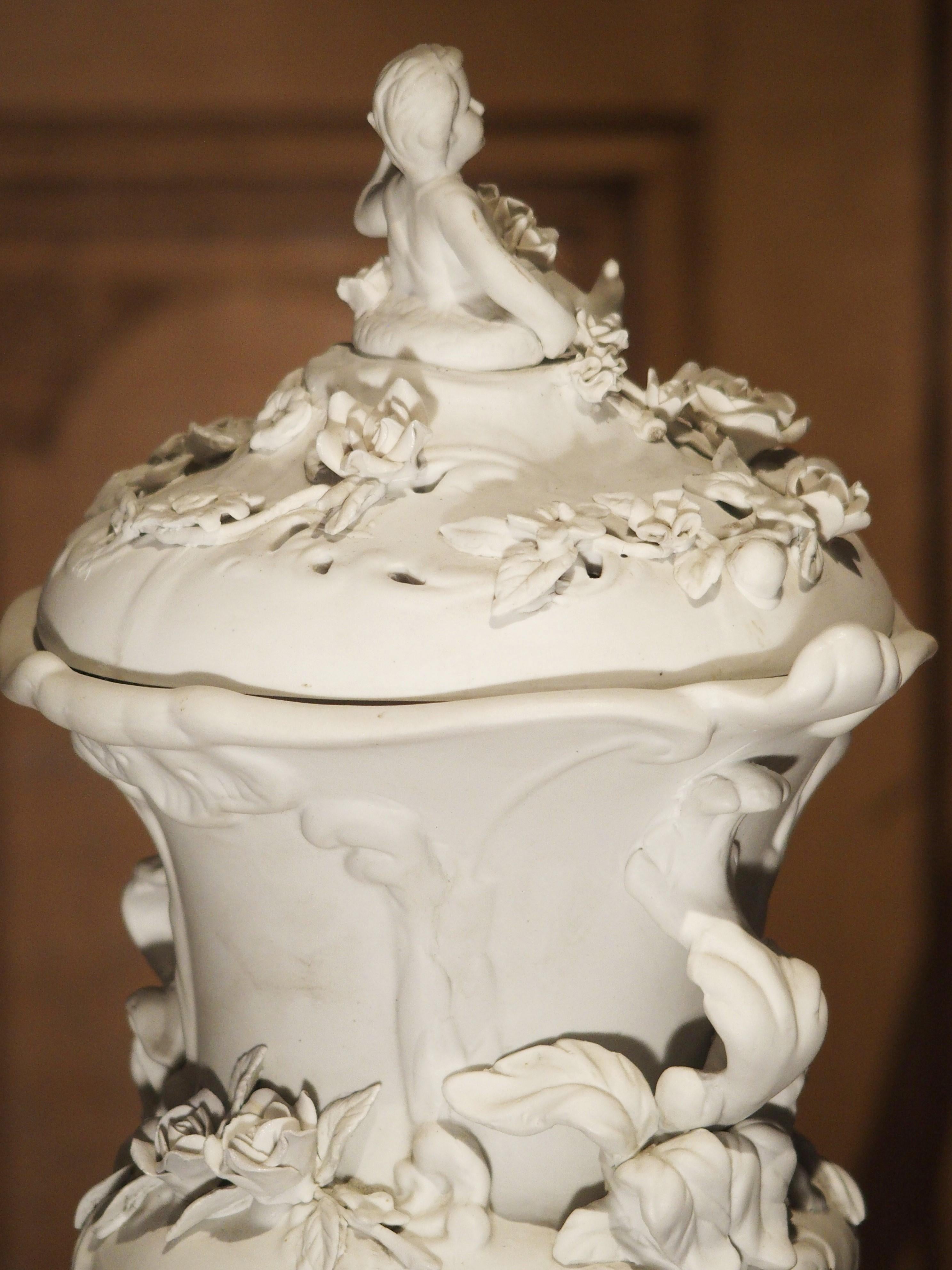 This magnificent antique French bisque piece is a tabletop lidded urn on a tripod pedestal. It features gros relief motifs of cherubs, foliate and floral vines, drapery with tassels, roses, cartouches and more. With a height of almost 23 inches,