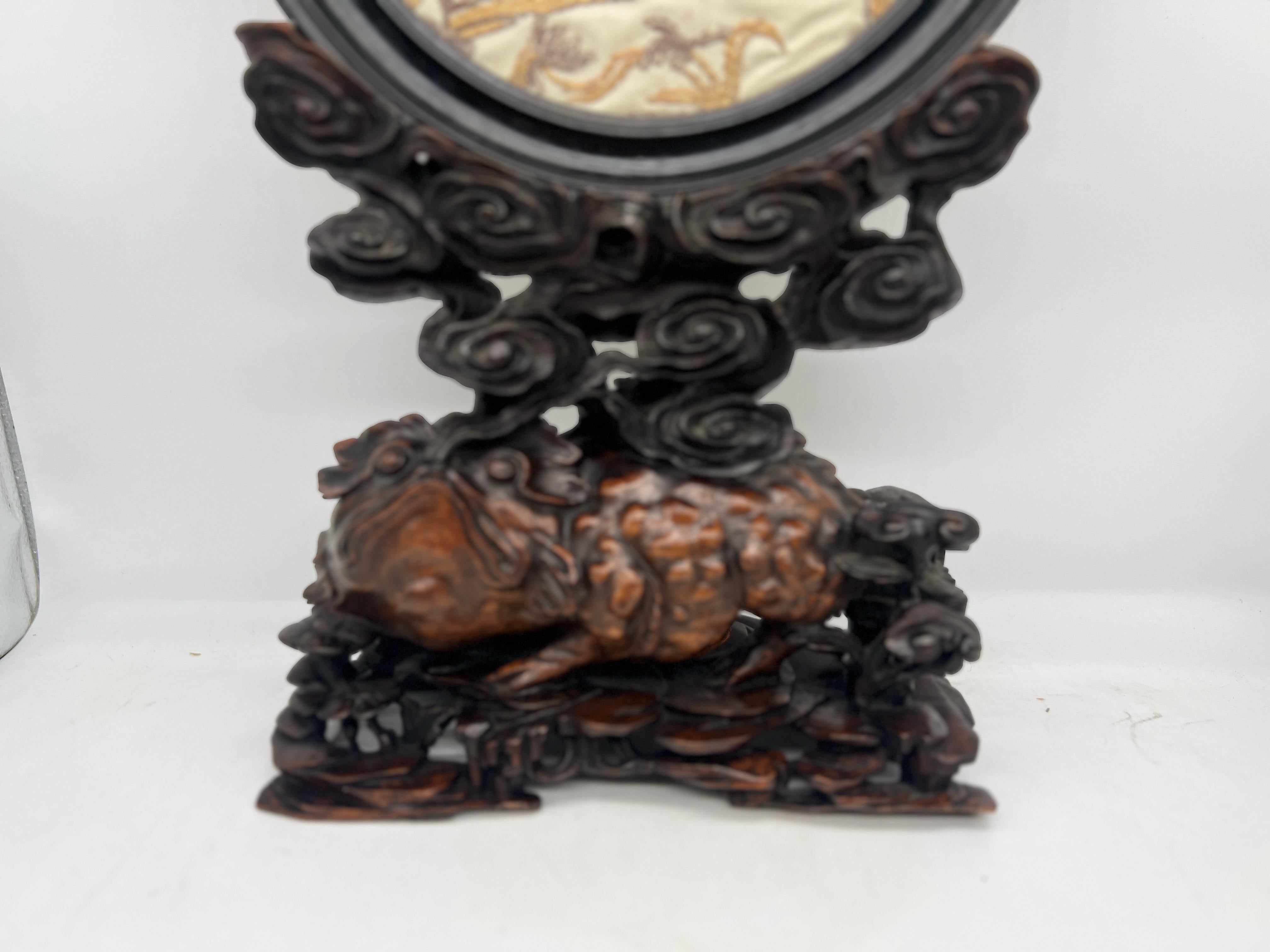 This finely crafted piece combines traditional Chinese textile artistry with intricate woodcarving. The central element is a circular silk embroidery, depicting a serene scene with figures set amidst a landscape of stylized trees and architectural