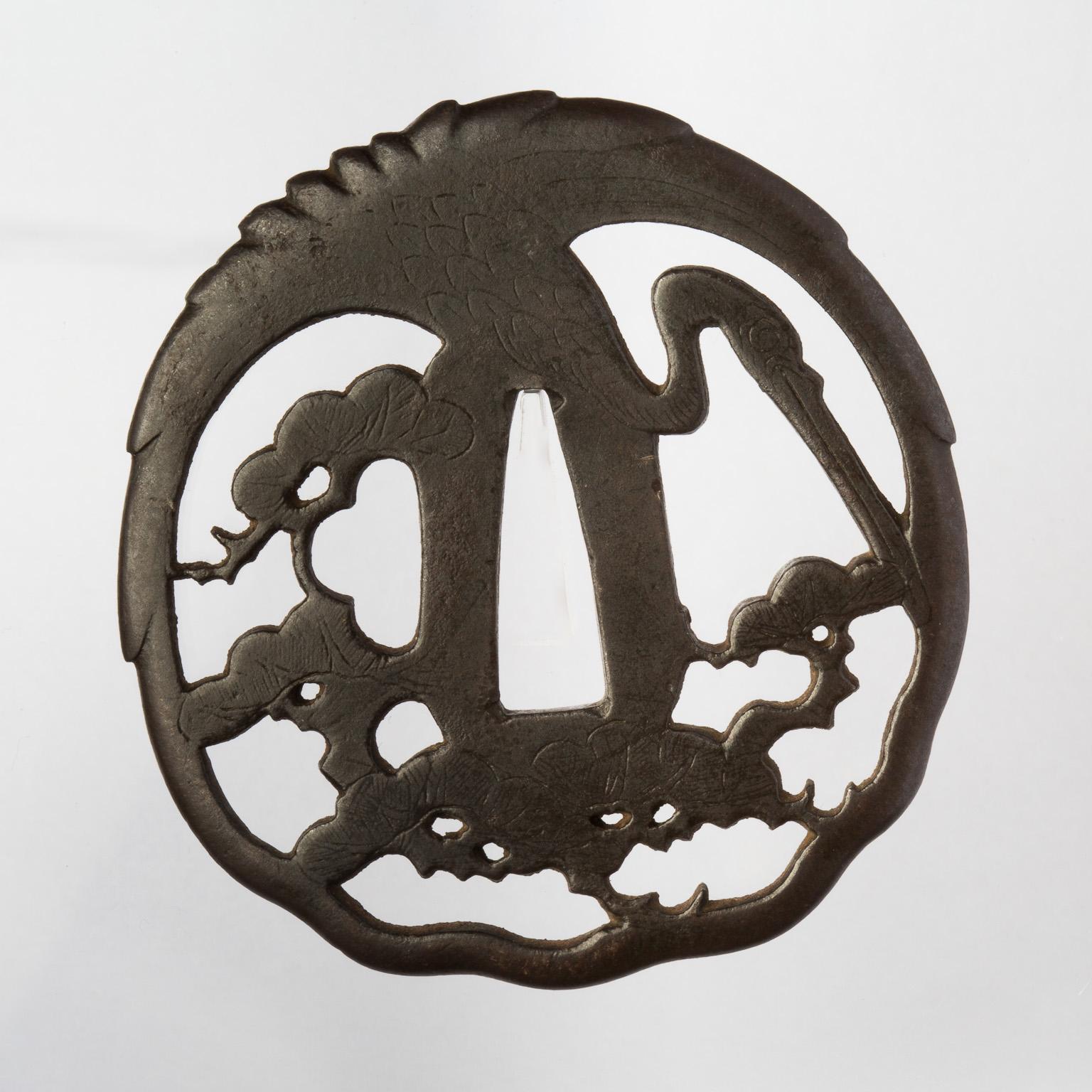 Maru-gata: 7.2 x 6.7 cm
Thickness: 4.2 mm
Higo Nishigaki tsuba representing a fine and elegantly stylized crane (tsuru) with outstretched wings, flying over some pines trees. The bird's wings form the upper edge of the tsuba itself.