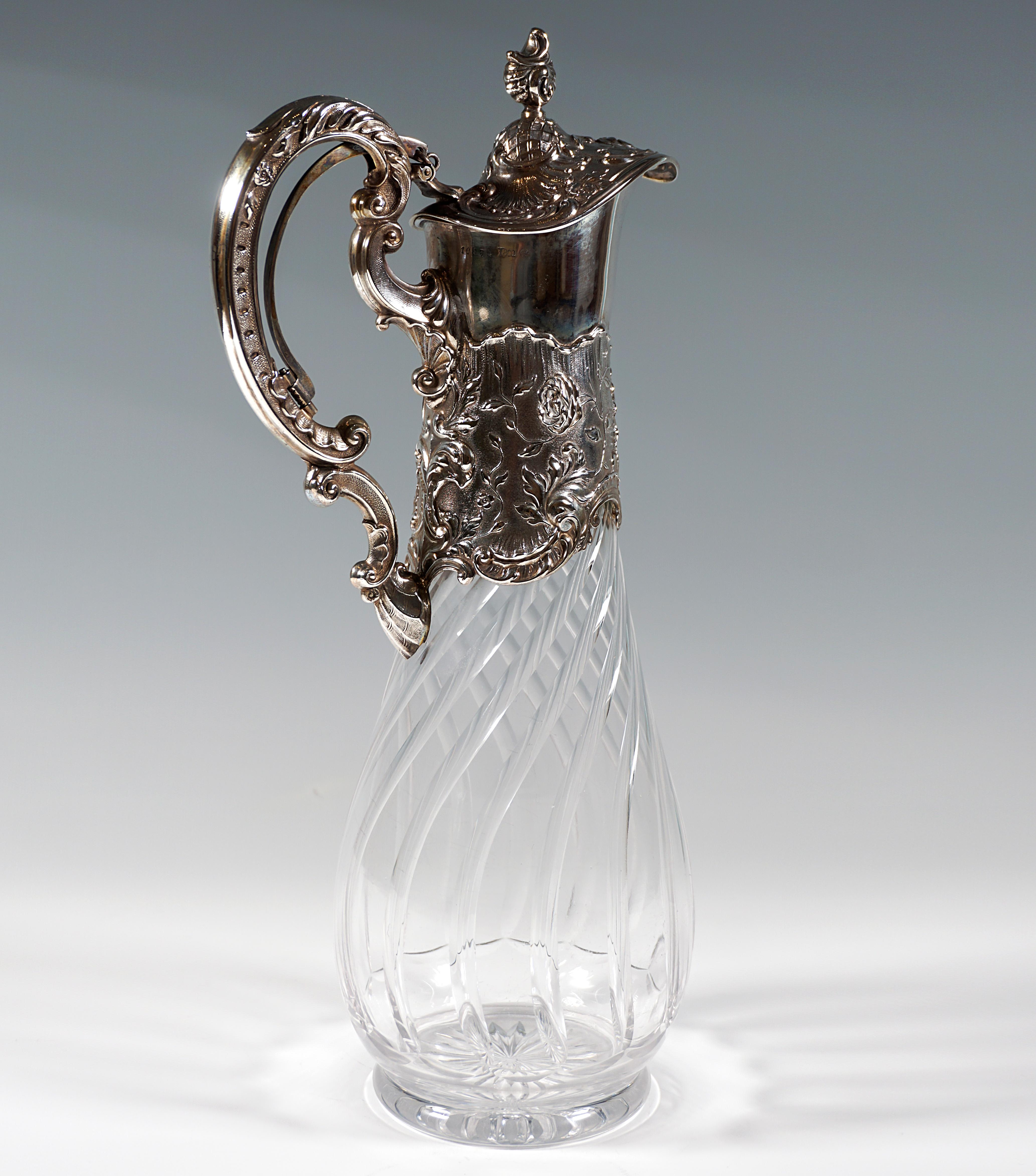 Carafe of clear glass in the shape of a slender drop on a stand ring, the wall with vertical, curved incisions, cut bottom star, the upper part of the glass body framed in silver mountings with a wide spout with lid and thumb rest, richly decorated