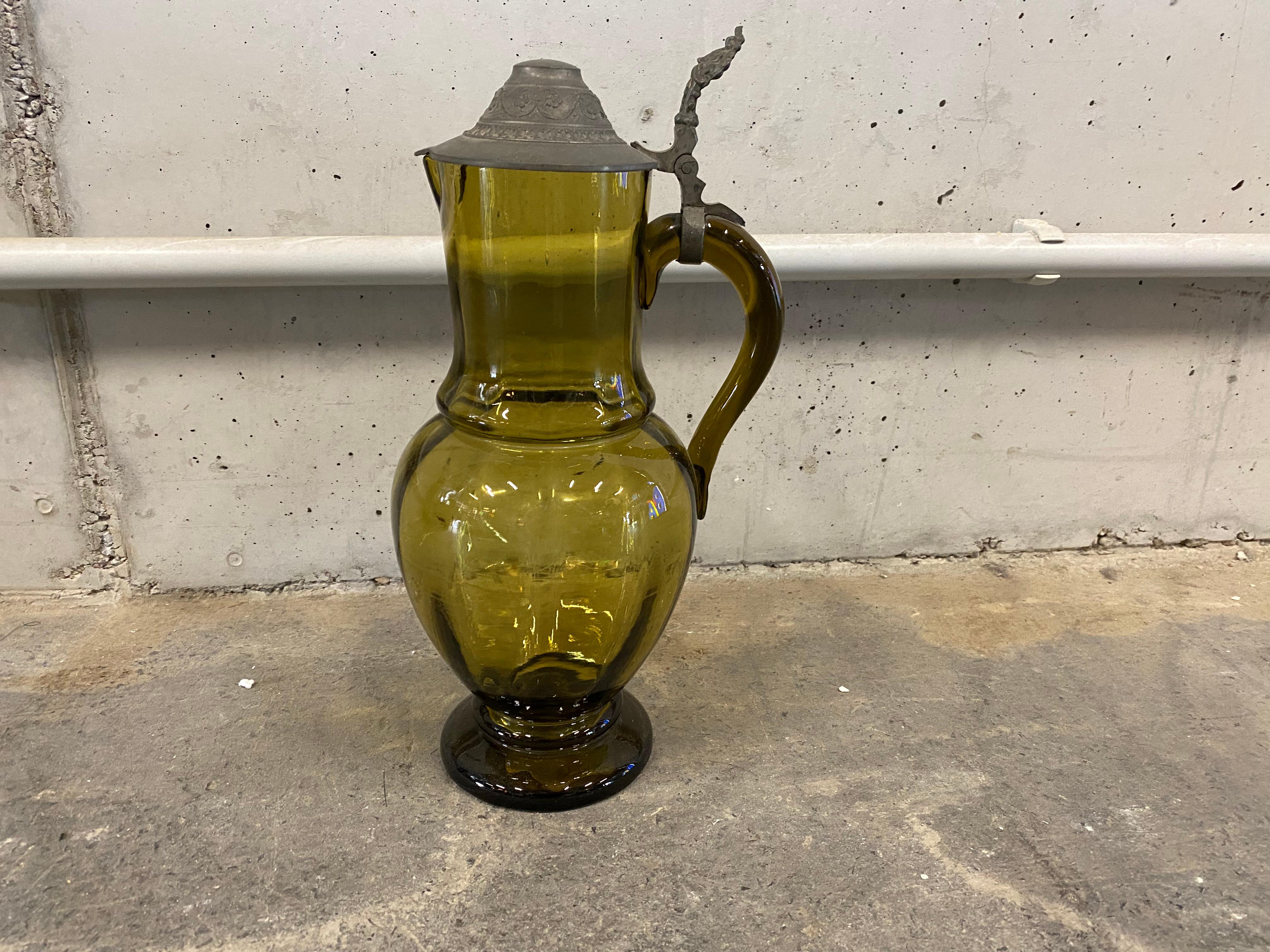 Glass jug with lid from the years around 1890. This beautiful mouth-blown jug made of olive green stained glass and lid made of tin is marked and belongs to the historicism. The ornate lid is attached to the handle of the pitcher and can be opened