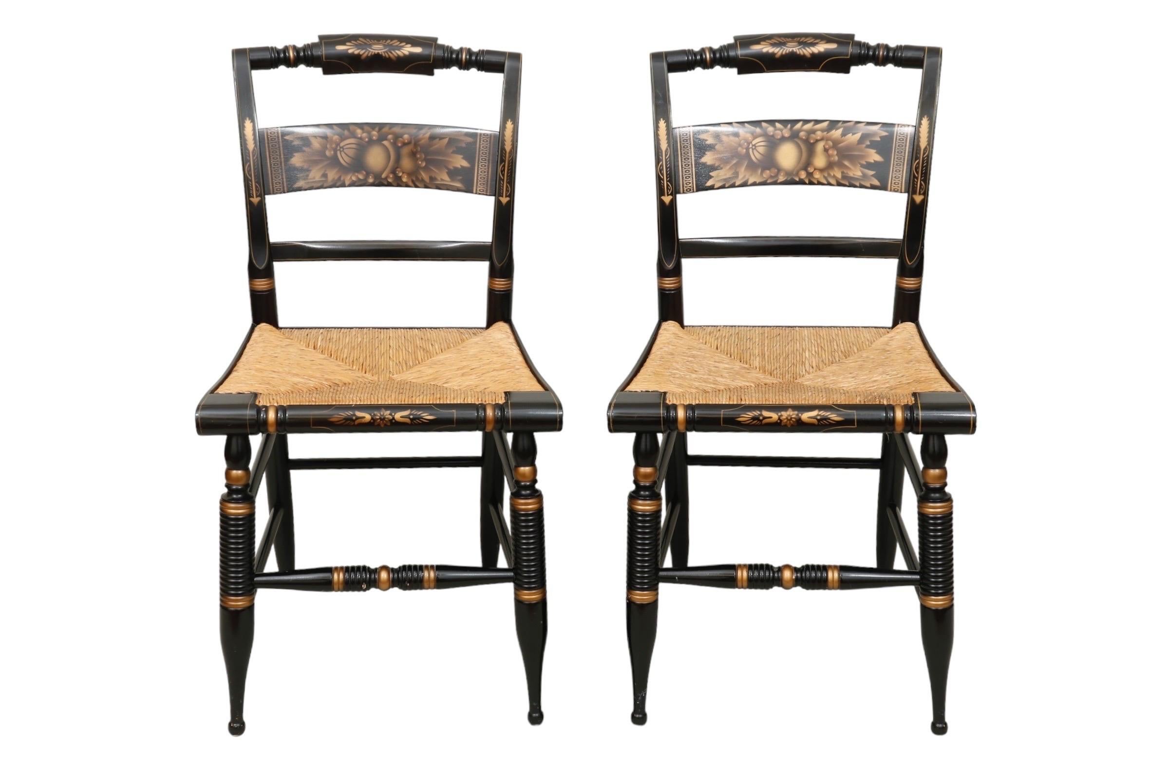 A pair of black and gold Lambert Hitchcock hand painted side chairs with traditional rush seats. Pillow box crest rails and flat back splats are stenciled with the harvest pattern. Detailed throughout with gold accents with a floral motif on the