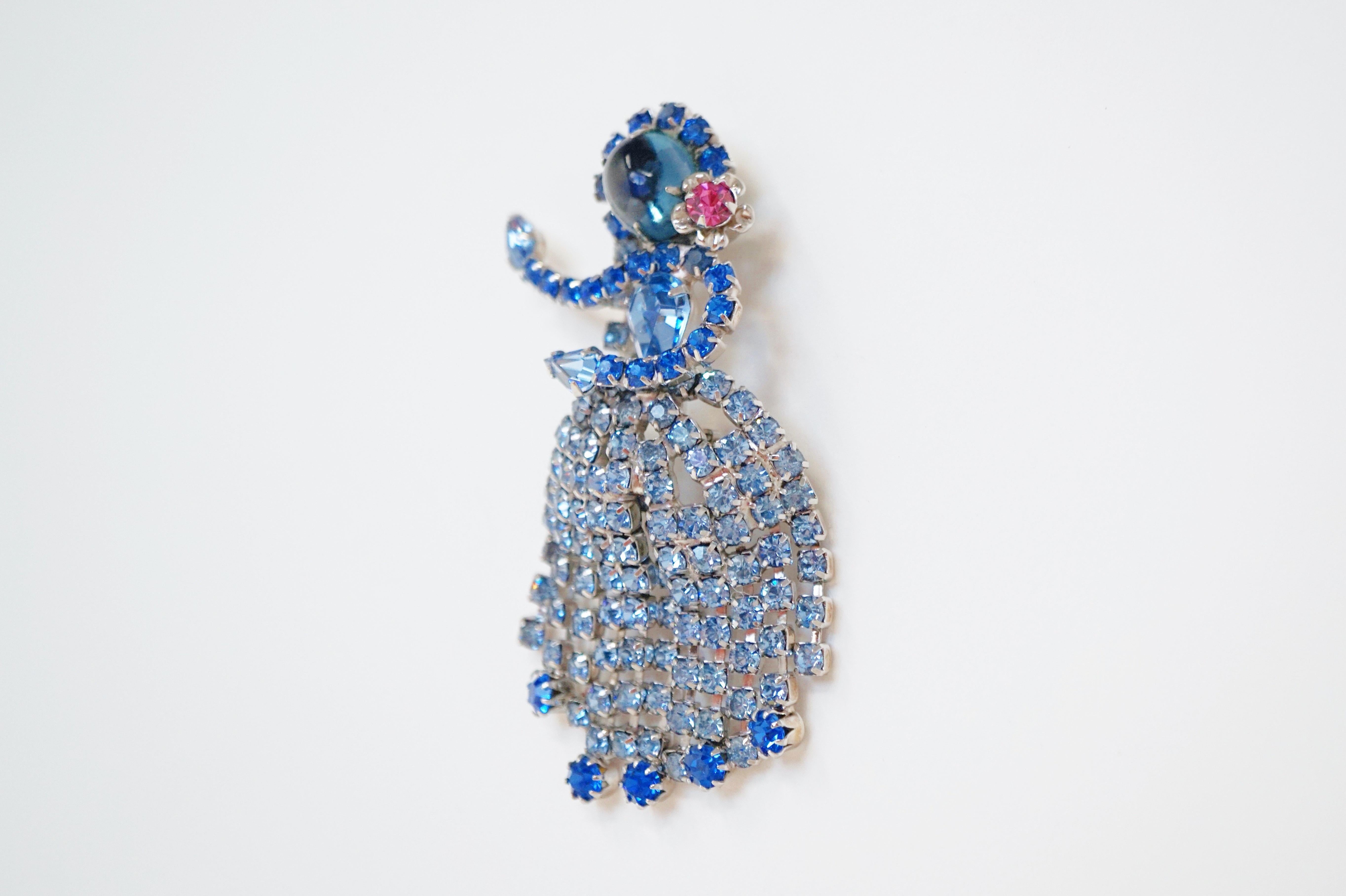 This gorgeous rhinestone Hula Dancer brooch by Hobé, circa 1965, features sky blue and azure rhinestones, a glass cabochon and a pink rhinestone flower accent with silver tone hardware. 
This brooch was manufactured during the mid-60's in several