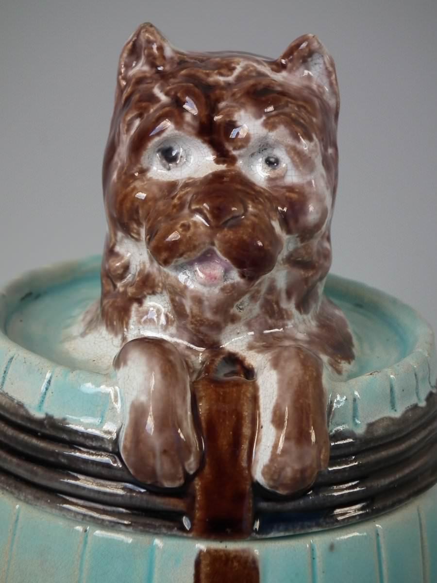 Majolica tobacco jar and cover which features a dog in a barrel. Turquoise ground version. Colouration: turquoise, brown, white, are predominant. Bears a pattern number, '12'.