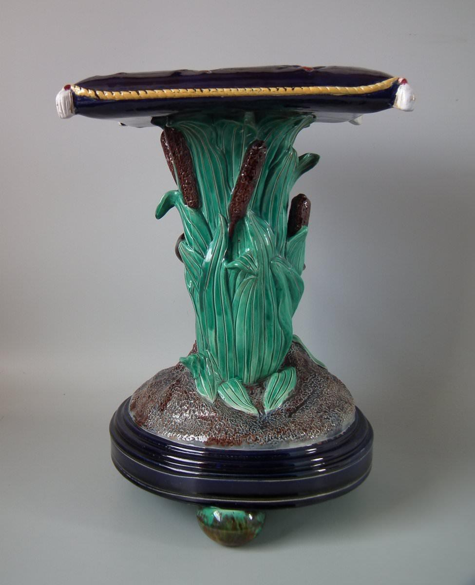 Holdcroft Majolica garden seat which features a boy sat in bull rushes (cattails), bird perched knee, supporting a red-buttoned cushion top. Coloration: cobalt blue, green, black, are predominant.