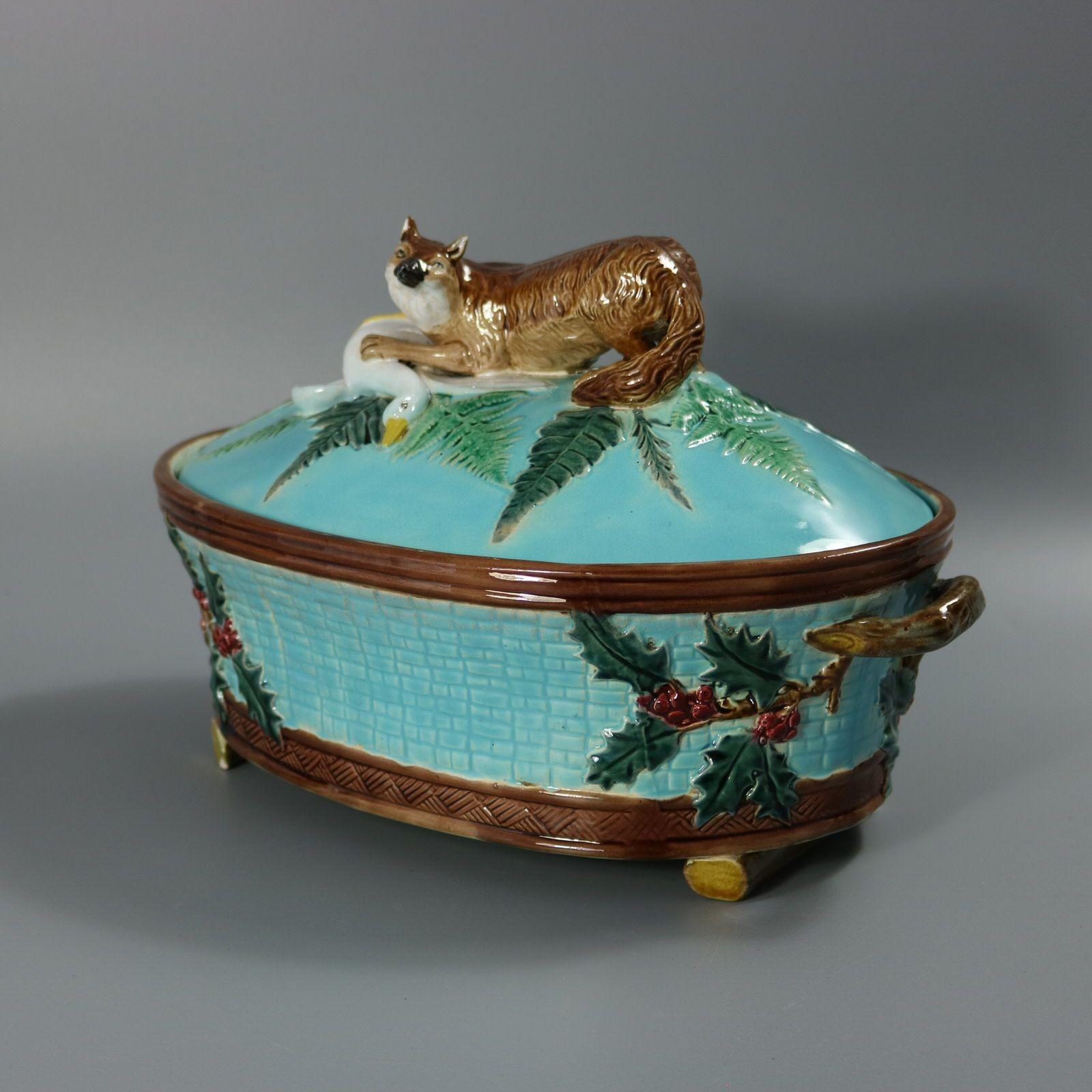 Holdcroft Majolica game pie dish which features a fox holding a goose down amongst ferns. Holly hanging off the branch-like handles. Turquoise ground version. Colouration: turquoise, green, brown, are predominant. Bears a pattern number, '+'.