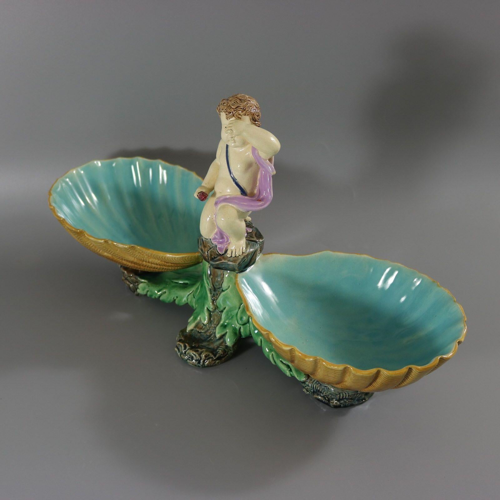 Le plat Holdcroft Majolica, qui représente un putto tenant une torche, flanqué de coquillages de chaque côté, est posé sur des rochers ornés d'algues. Coloration : jaune, vert, turquoise, sont prédominants.