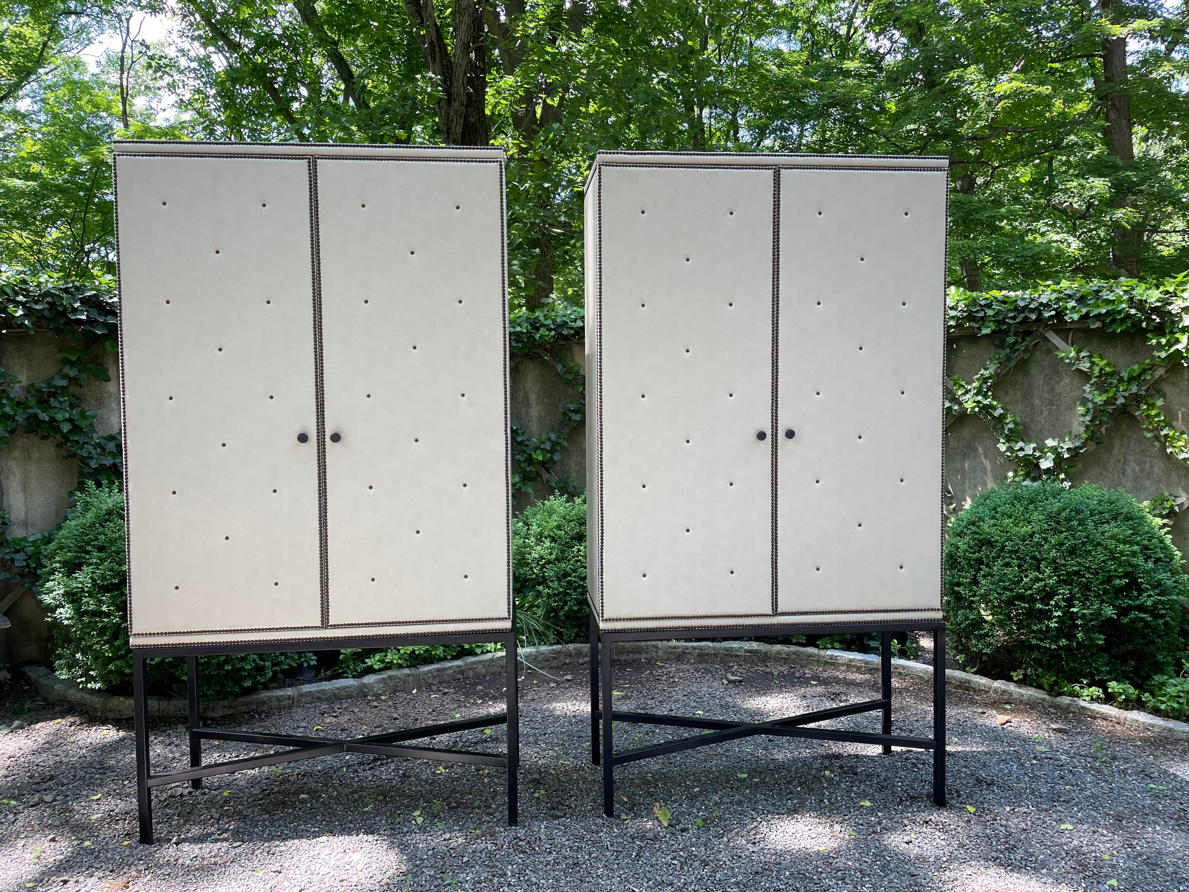 Very glamorous pair of custom media cabinets upholstered in padded beige leather and finished with bronze nailheads. Each cabinet sits on criss crossed steel bases. The backs have circles and rectangles cut to accomodate cords and wires. Upper shelf