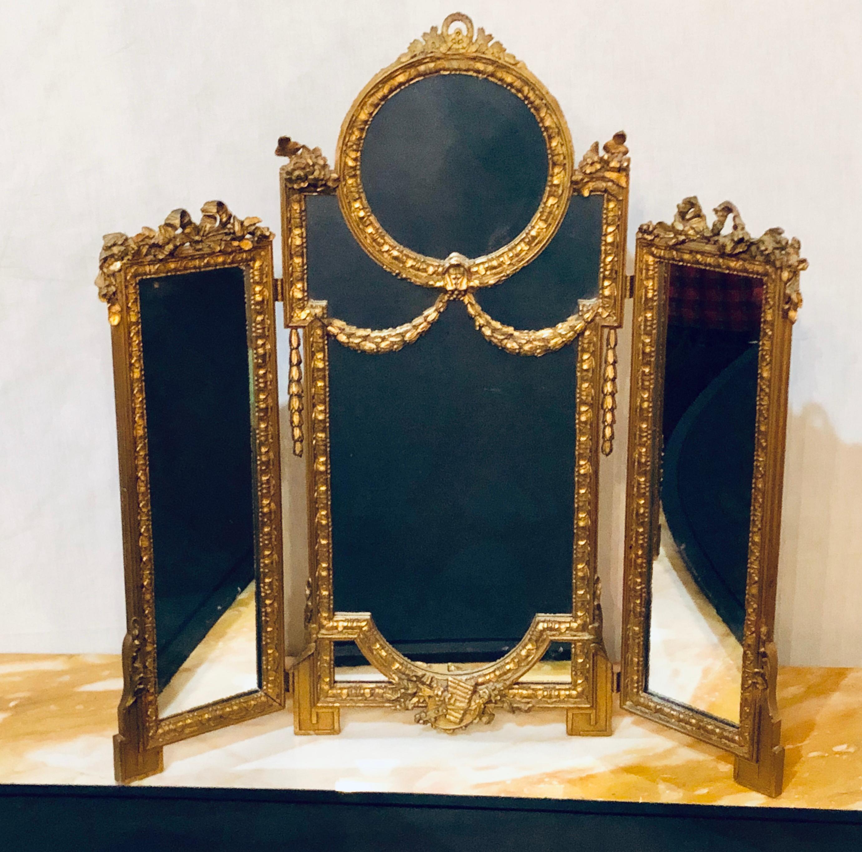 Hollywood Regency 1940s Louis XVI Style Gilt wood Tri-fold Vanity or Table Mirror. This small table or vanity mirror is simply as sweet as can be. The Louis XVI carved gilt wooden frame having the standard bow and arrow with tassel carvings one
