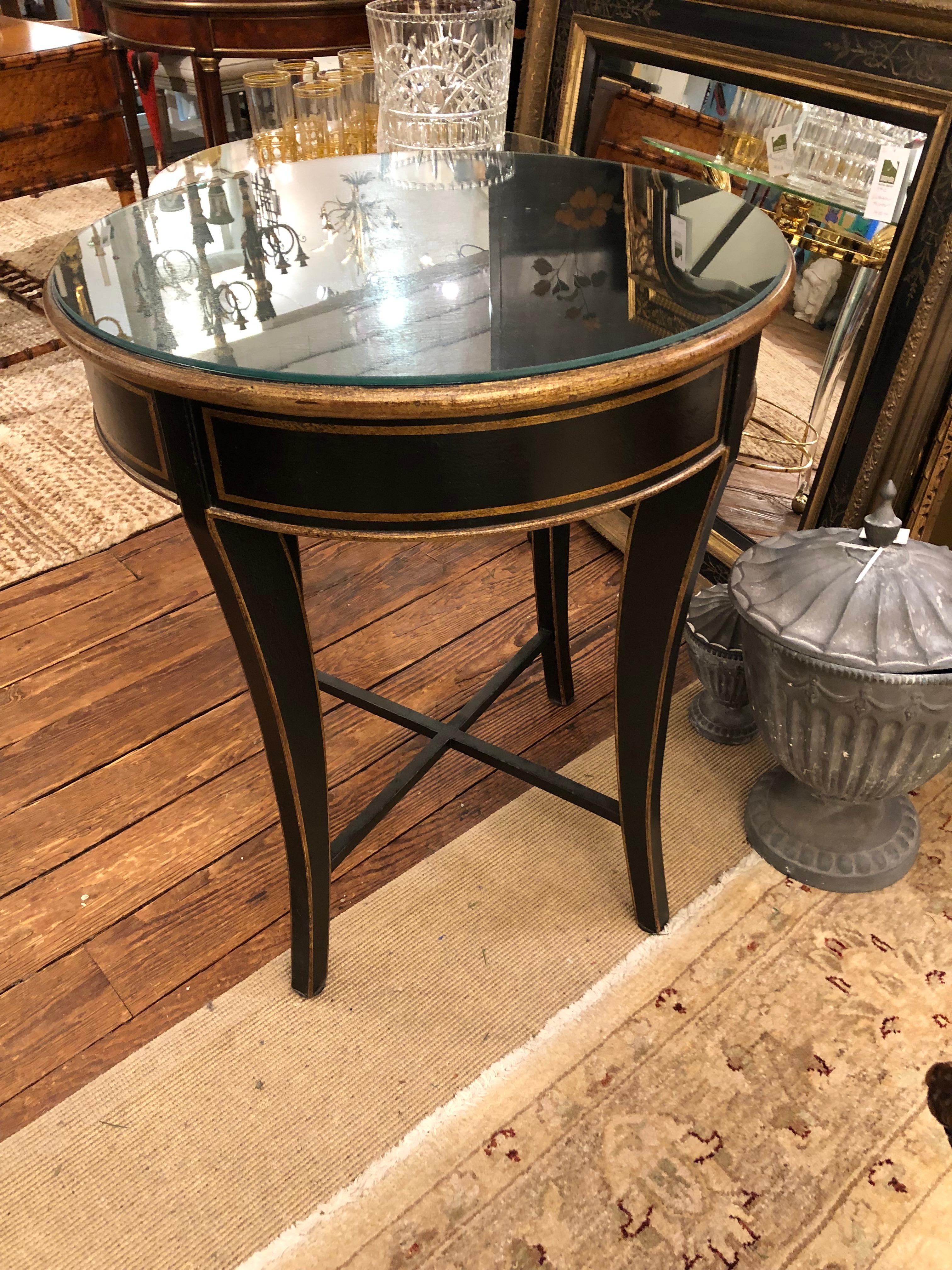 Transitional Hollywood Regency style round center or side table in black and gold, having tapered slightly splayed legs and handsome stretcher. Comes with heavy piece of protective glass on top.