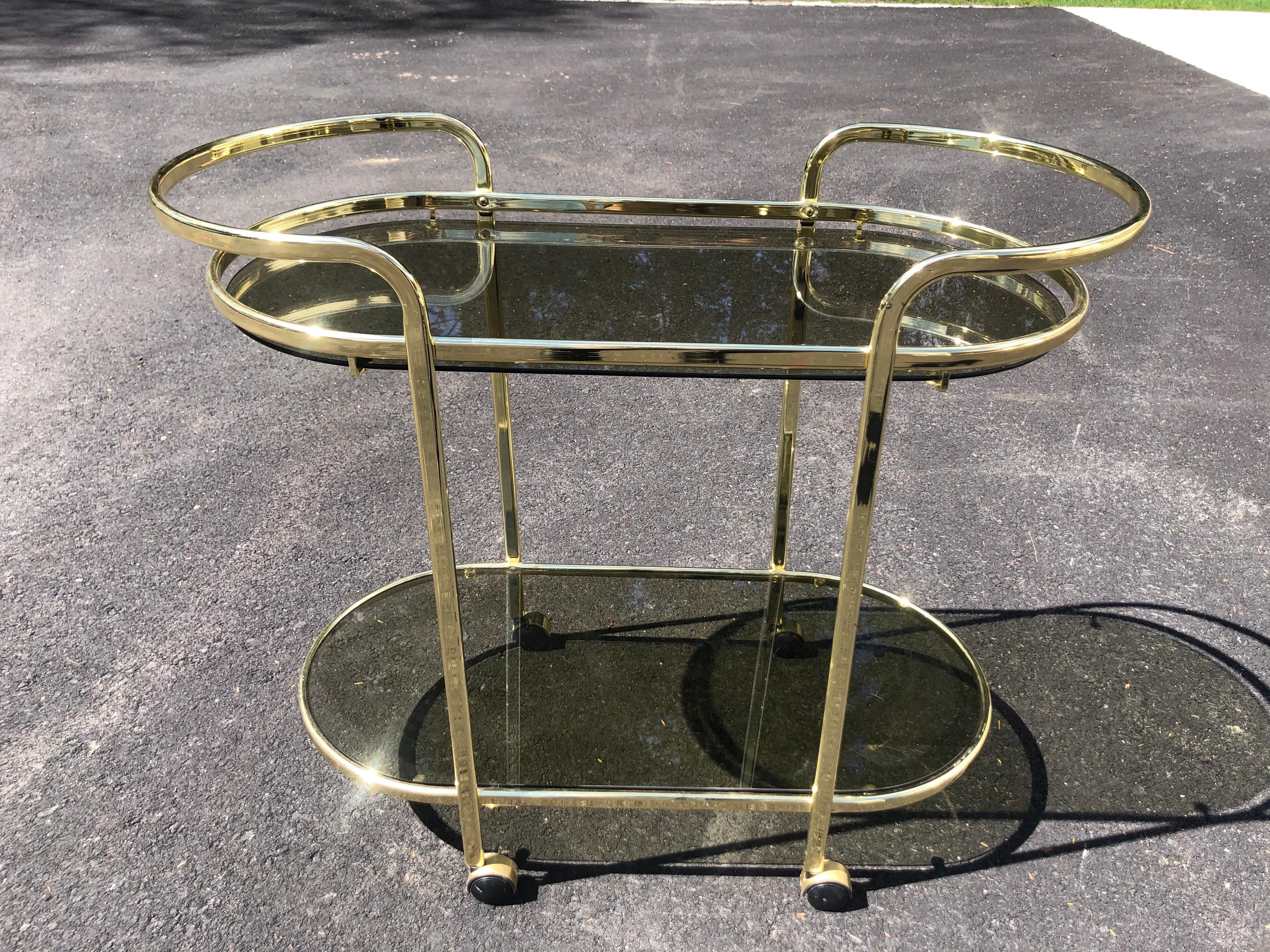 Hollywood Regency brass and smoked glass bar cart. Sleek and sexy defines this minimalist cart. Perfect in that upscale home with chic design. Two tiered for plenty of storage. Wheel from room to room for your convenience.