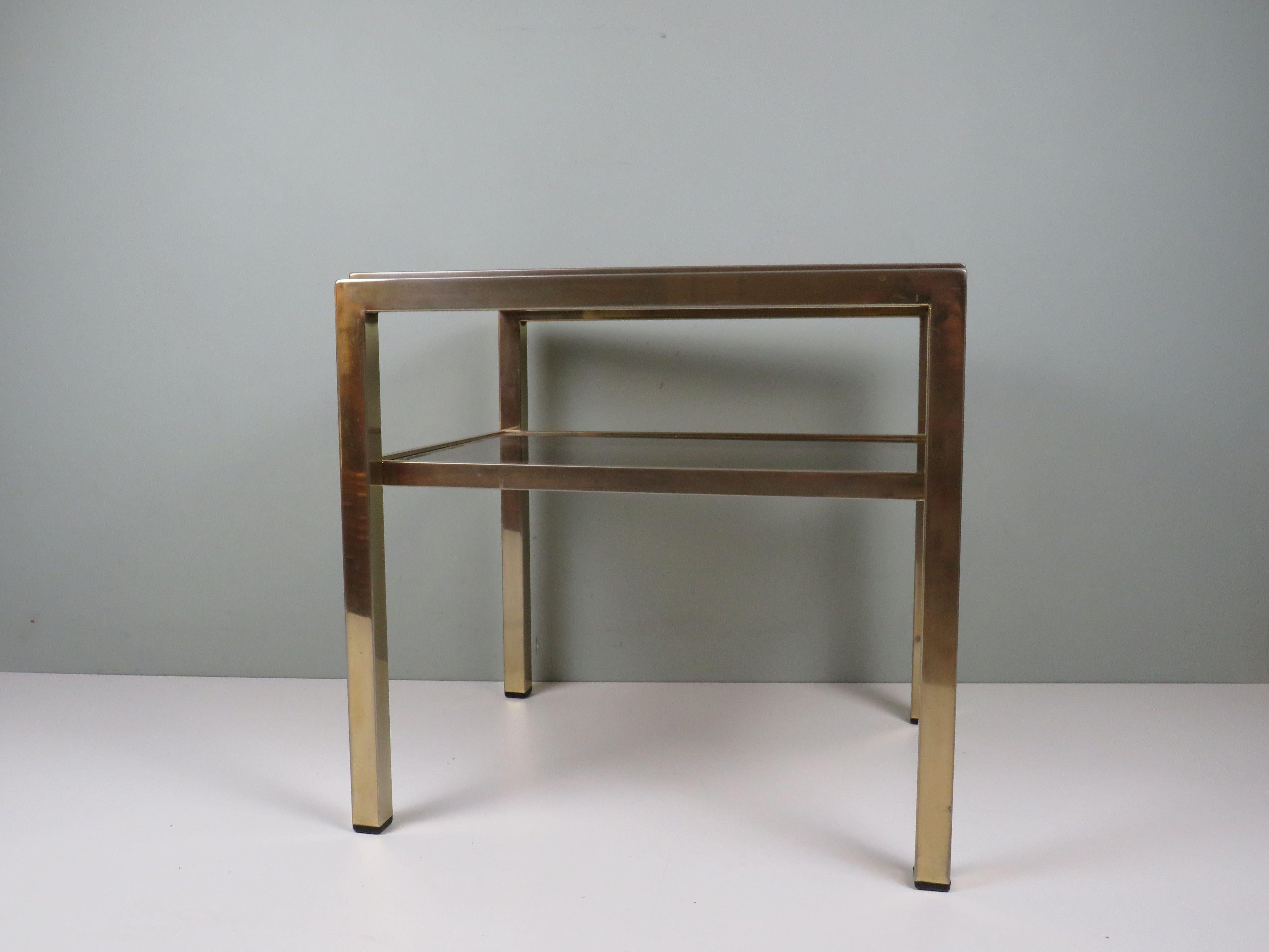 Table d'appoint sophistiquée de style Hollywood Regency, conçue et produite par De Knudt, Belgique, vers 1970. Le cadre en laiton massif présente des lignes architecturales nettes, complétées par deux niveaux en verre fumé qui apportent de la