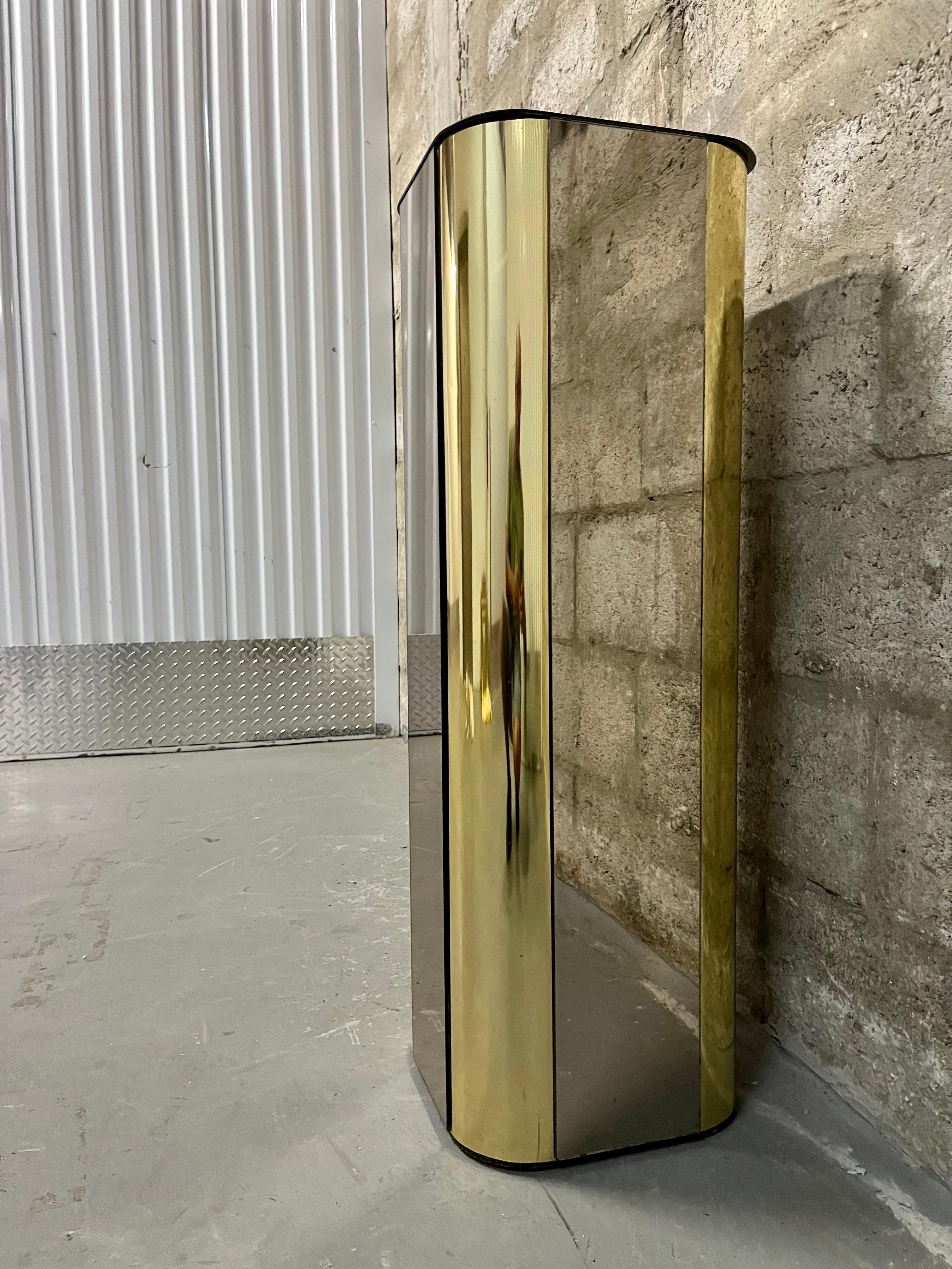 Vintage Hollywood Regency Brass and Smoked Mirror Pedestal in the Curtis Jere's Style. Circa 1970s 
Features a brass rounded edged with smoked mirror side panels and smoked mirror with rounded corners top. 
In good original condition with minor