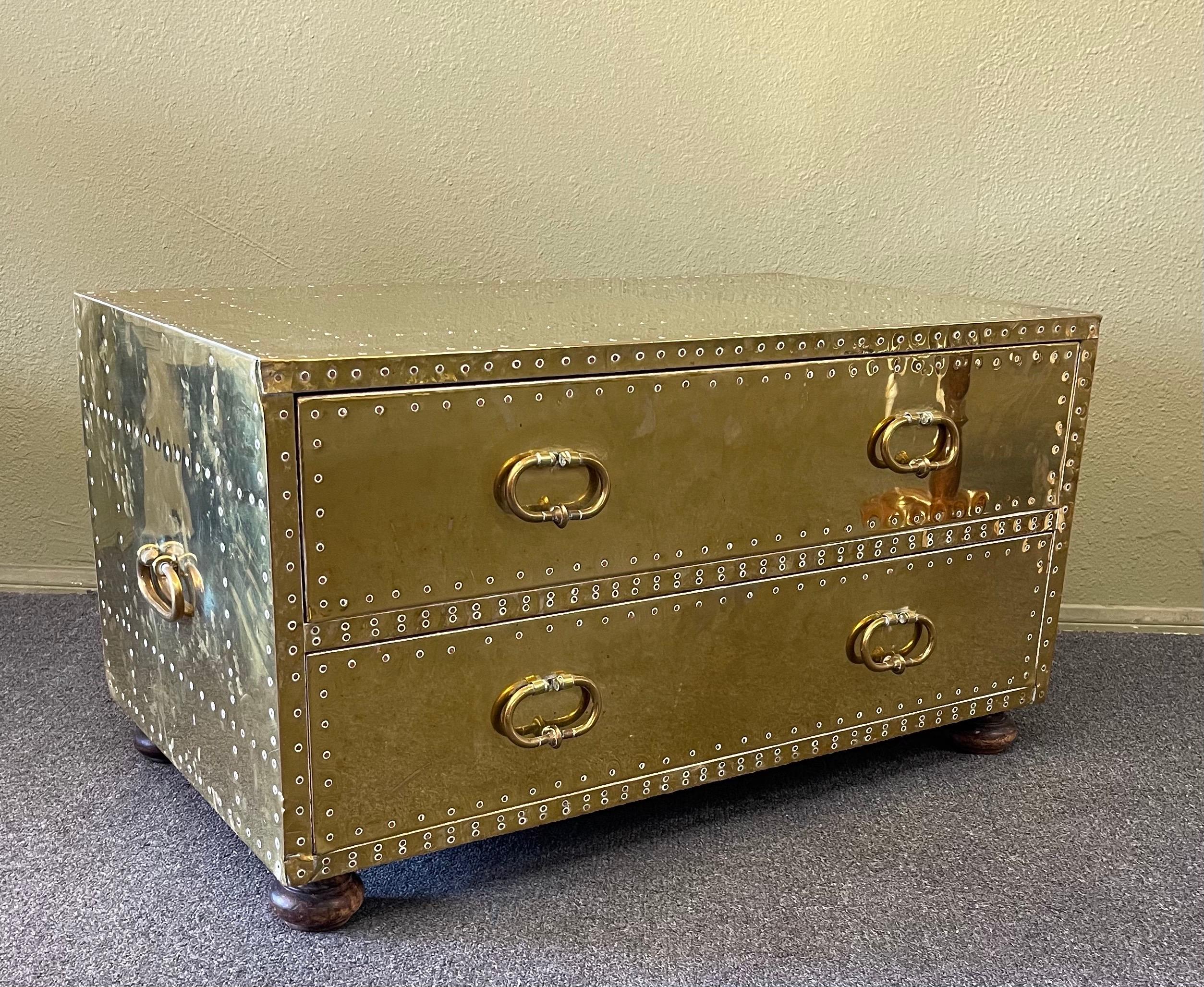 A very nice Hollywood Regency brass clad two drawer chest by Sarreid LTD of Spain, circa 1970s. The chest is constructed of solid wood with dovetailed drawers covered in brass clad with hammered brass nails and wood bun feet. It is in good vintage