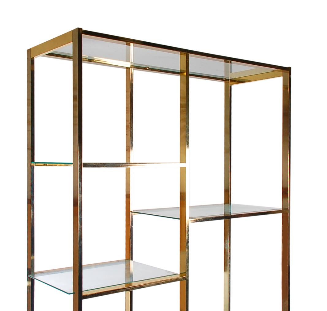 A Classic asymmetrical design wall unit in the manner of Milo Baughman. It features brass framing and clear glass shelving.