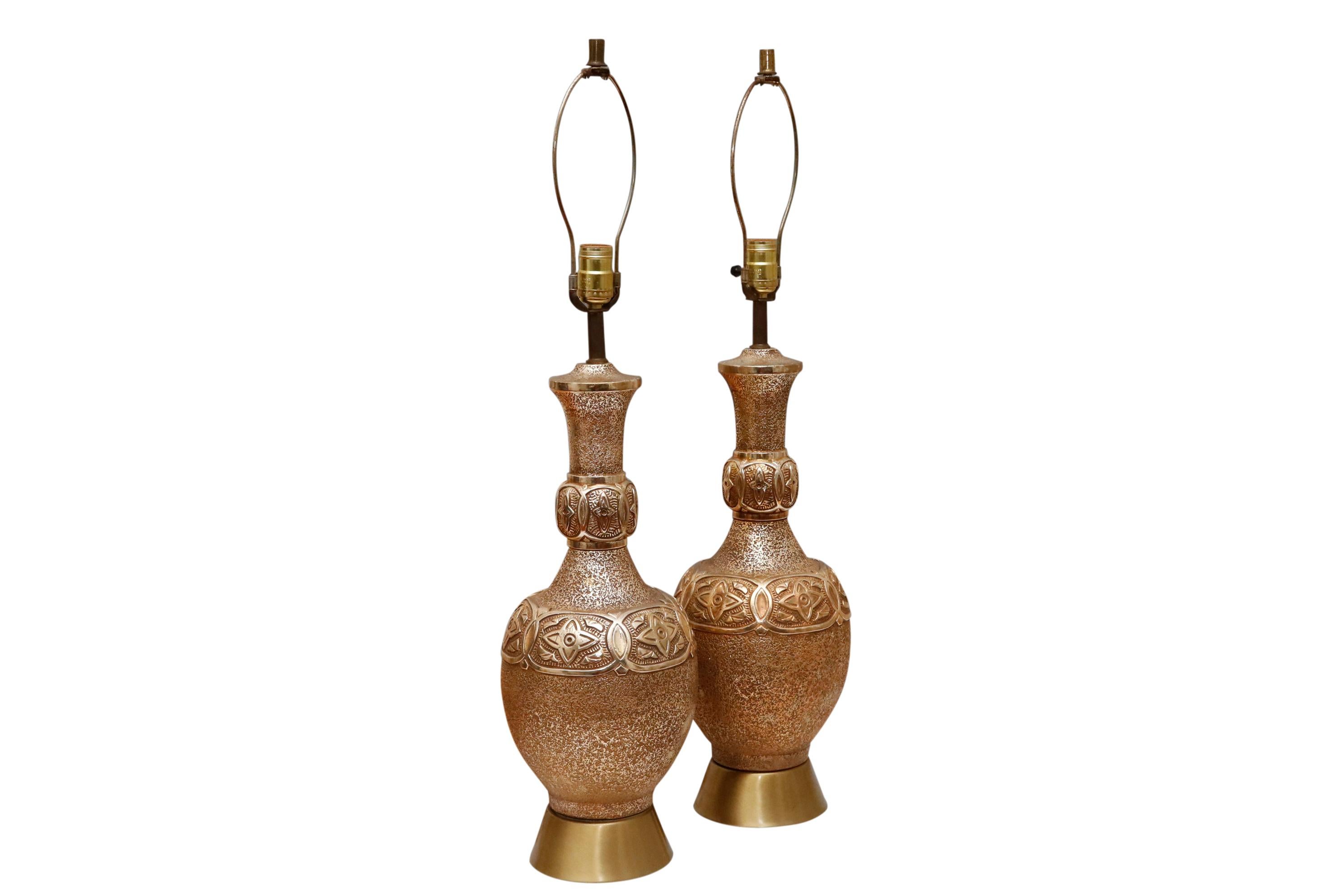 A pair of Hollywood Regency style brass table lamps. Decorated with a textured finish in a metallic gold color. Urns have an overlapping cartouche motif with a four pointed star at the shoulder and on the neck. Thick polished brass bases flare out