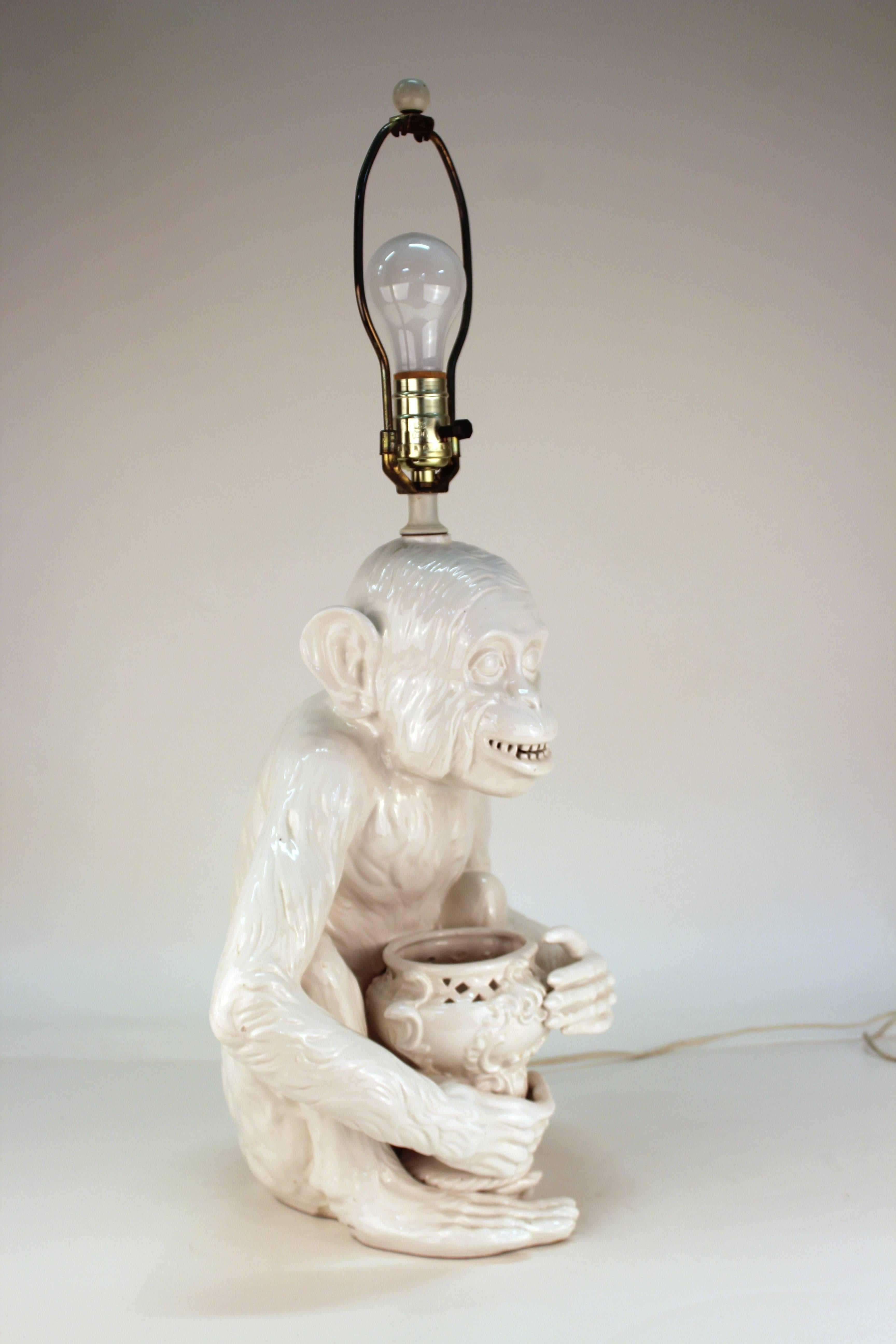 A Hollywood regency ceramic table lamp in shape of a seated grinning monkey holding an urn. The piece is in good vintage condition with age-appropriate wear.