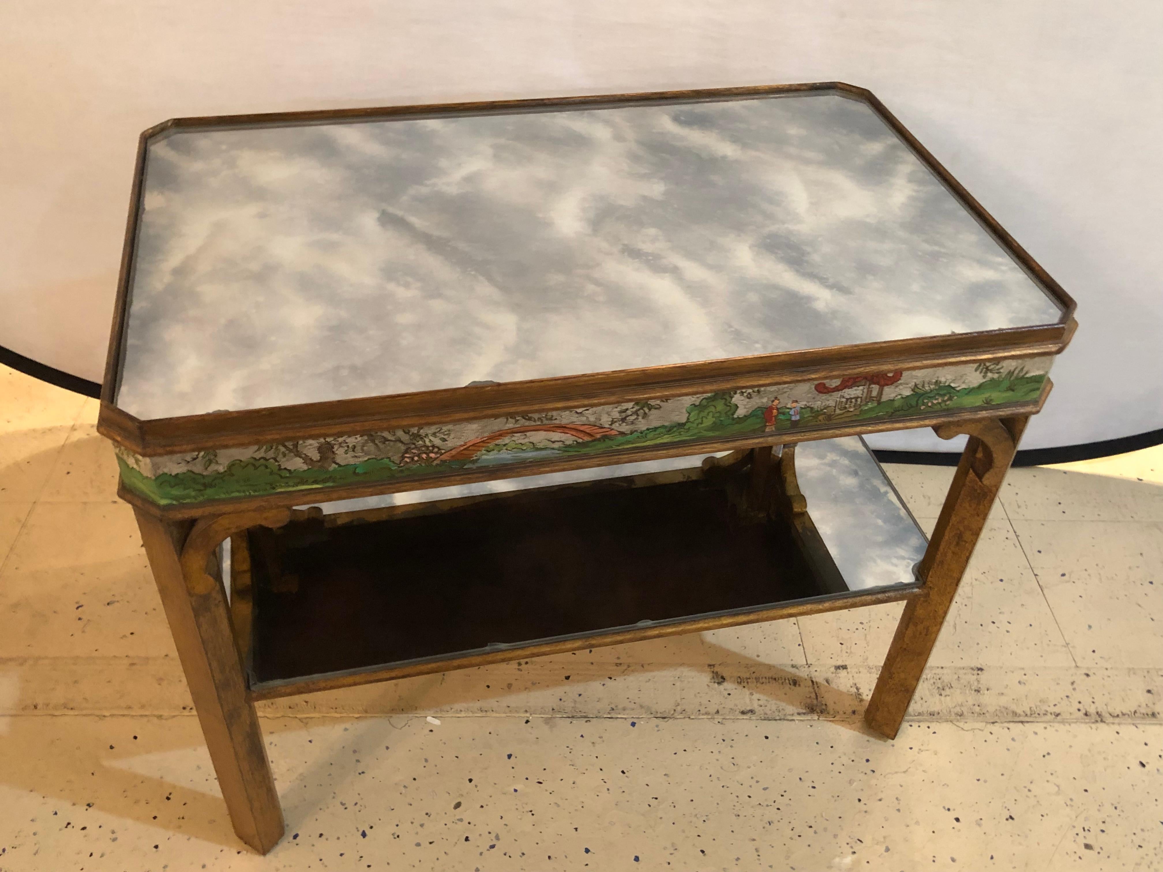 Hollywood Regency coffee table with poly-chromed mirrored scenes in chinoiserie. This stunning and very sweet coffee or low table has one lower distressed mirror shelf with a distressed mirror top supported by a gilt gold frame flanking a mirrored