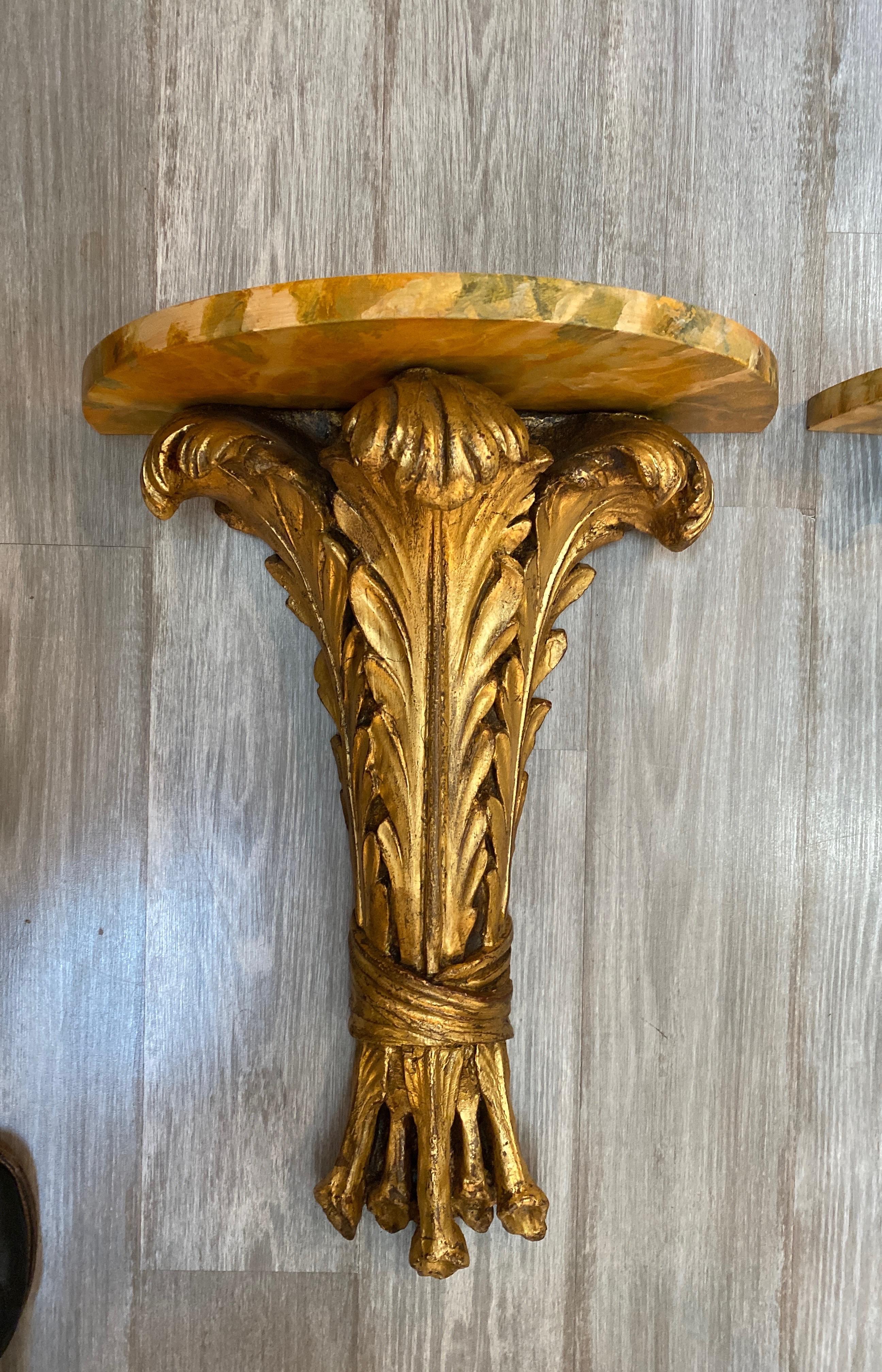 A pair of chic high style Hollywood Regency gilt and faux painted marble demi lune wall hanging tables. The acanthus leaf bases in solid dense plaster with gilt finish. The Demilune wood attached tops in a faux sienna marble. The shelves mount to