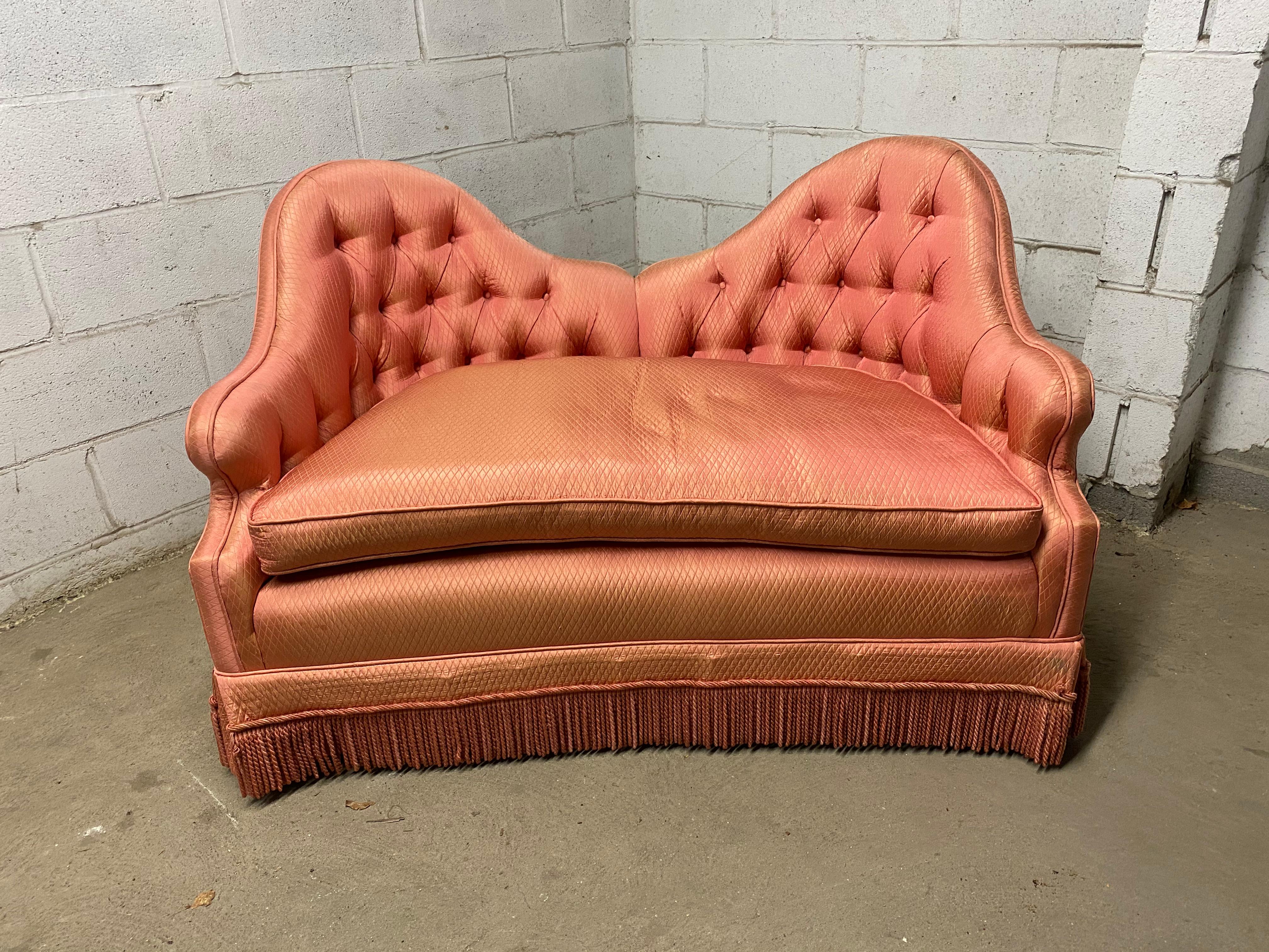 Hollywood Regency style tufted camel back loveseat. Visually dramatic and lovely. Amazing lines and details. Circa 1970. Upholstered in a silk salmon fabric. Fringe skirt. 

Structurally sound and sturdy frame. The fabric has signs of fading due