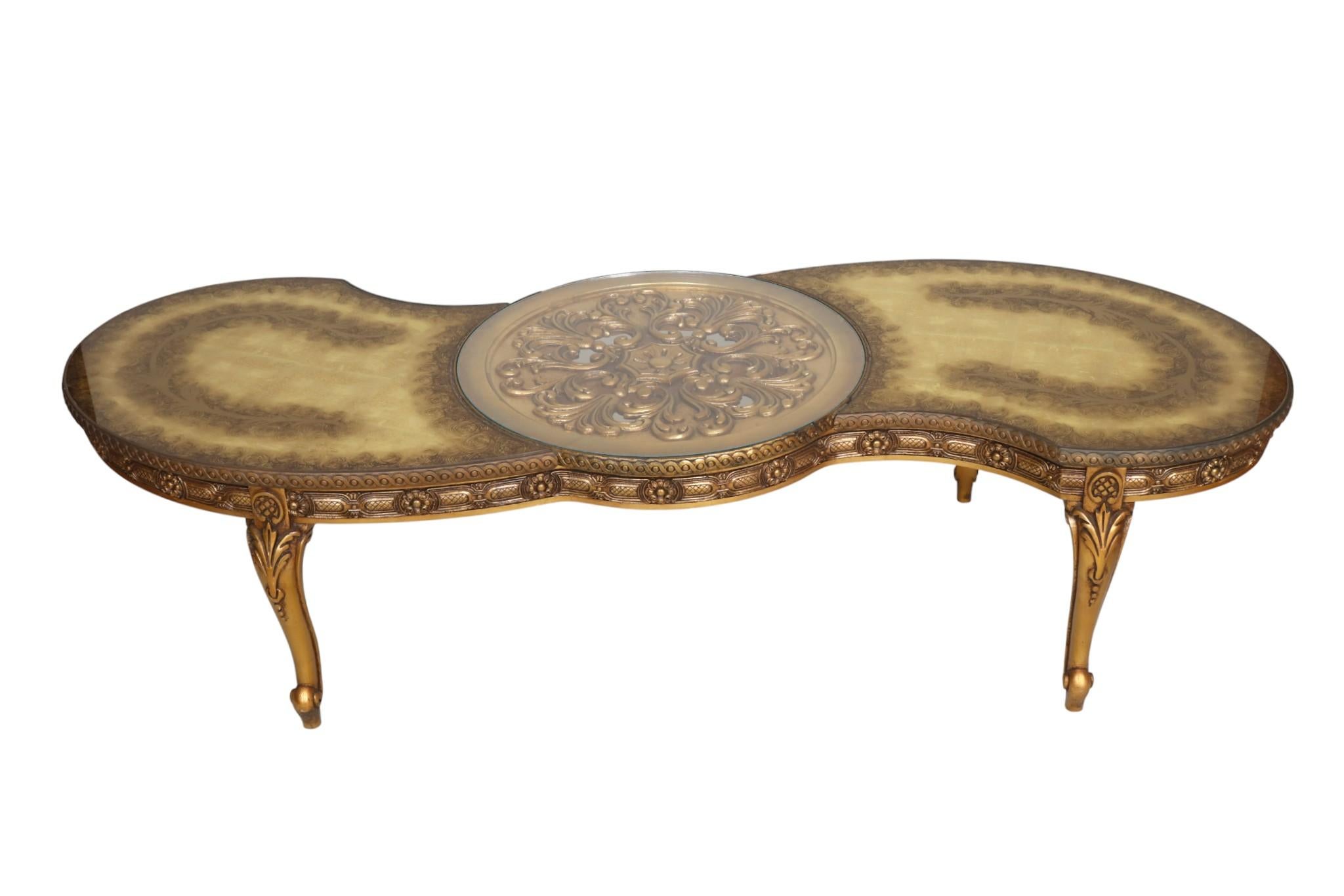 Table basse en bois doré et églomisé de style Régence hollywoodienne. Un médaillon central ajouré et sculpté est surmonté d'un plateau de verre rond amovible. De chaque côté, des plateaux de verre fixes sont peints en dessous avec une finition