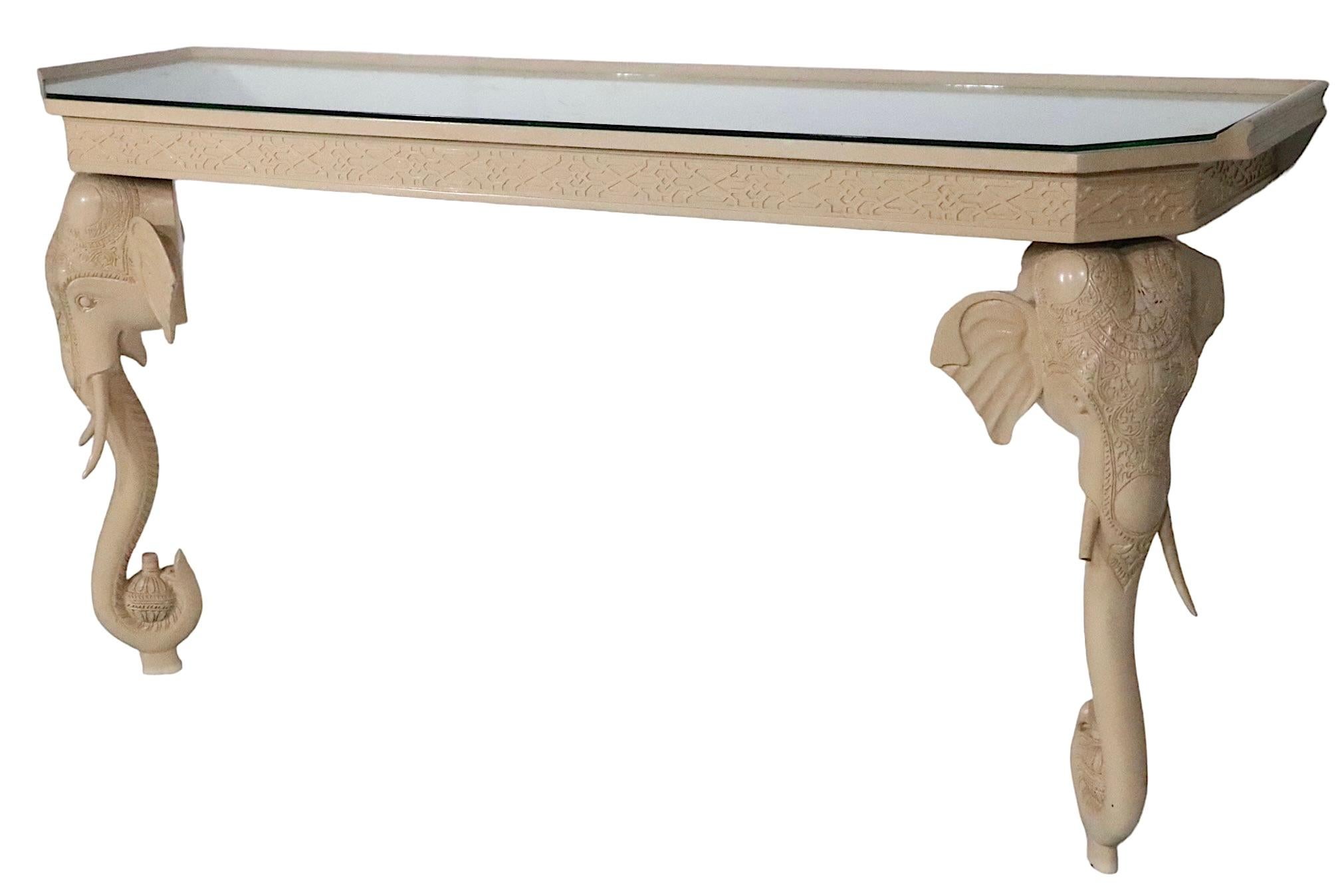 Chic Hollywood Regency console table having stylized elephant head, and trunk form legs. The table is of carved wood, in original cream, or off white, paint finish. This example is in good condition, it is structurally sound and sturdy, the paint