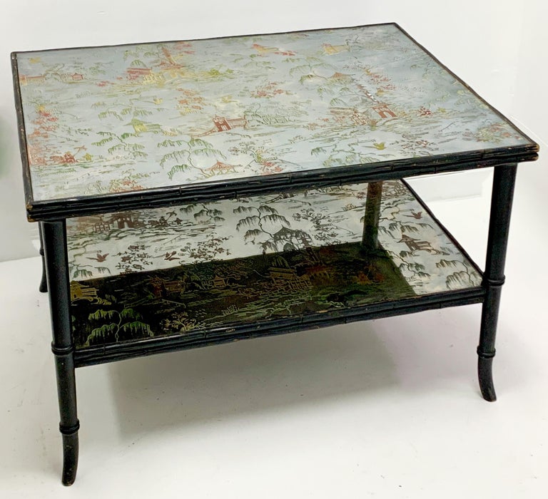 This is a faux bamboo two tiered coffee table with two tiered églomisé chinoiserie scenes on mirrored glass. A rare bird! The frame is a black lacquer carved wood. It is a midcentury piece and most likely Italian.