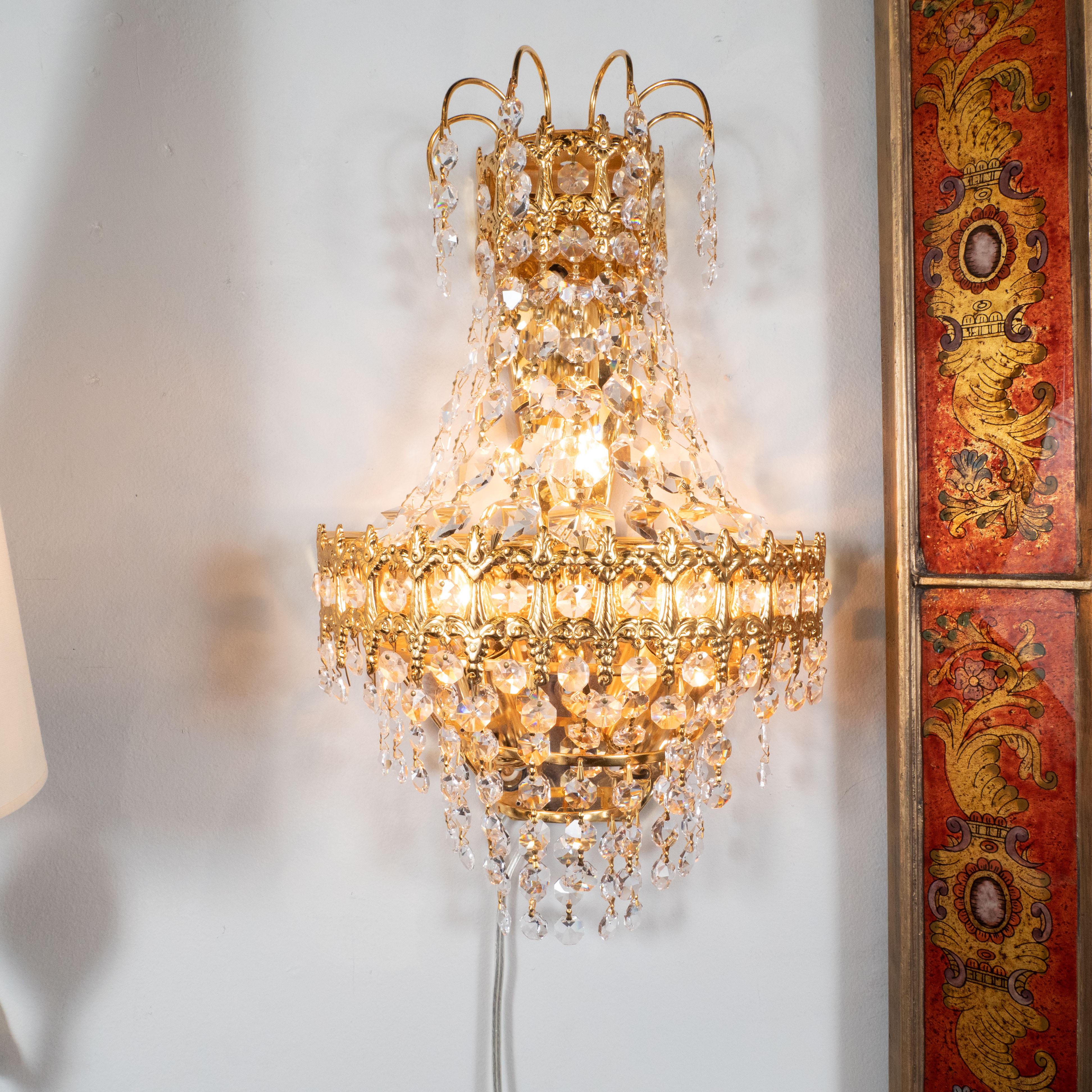 This glamorous pair of Hollywood Regency sconces were realized in the United States, circa 1945. They feature two demilune gold plated tiers with neoclassical detailing connected by strands of octagonal faceted crystal pendants. Crystals of the same