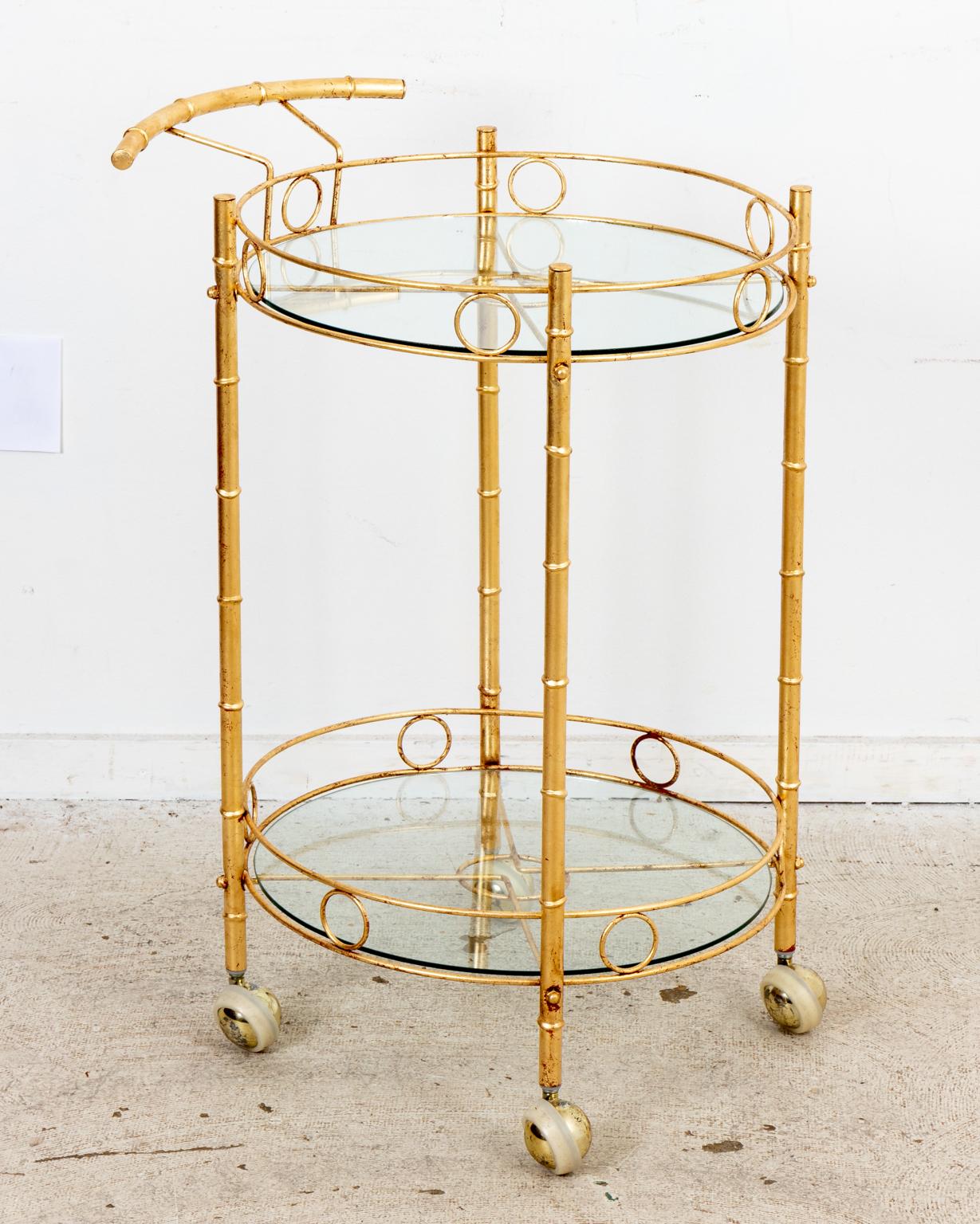 Circa 1960-1970s Hollywood Regency faux bamboo gilt metal round drink cart on ball shaped casters with angled metal detail. The drink cart also features two tiers with round glass inserts and disk trim gallery. Made in the United States. Please note