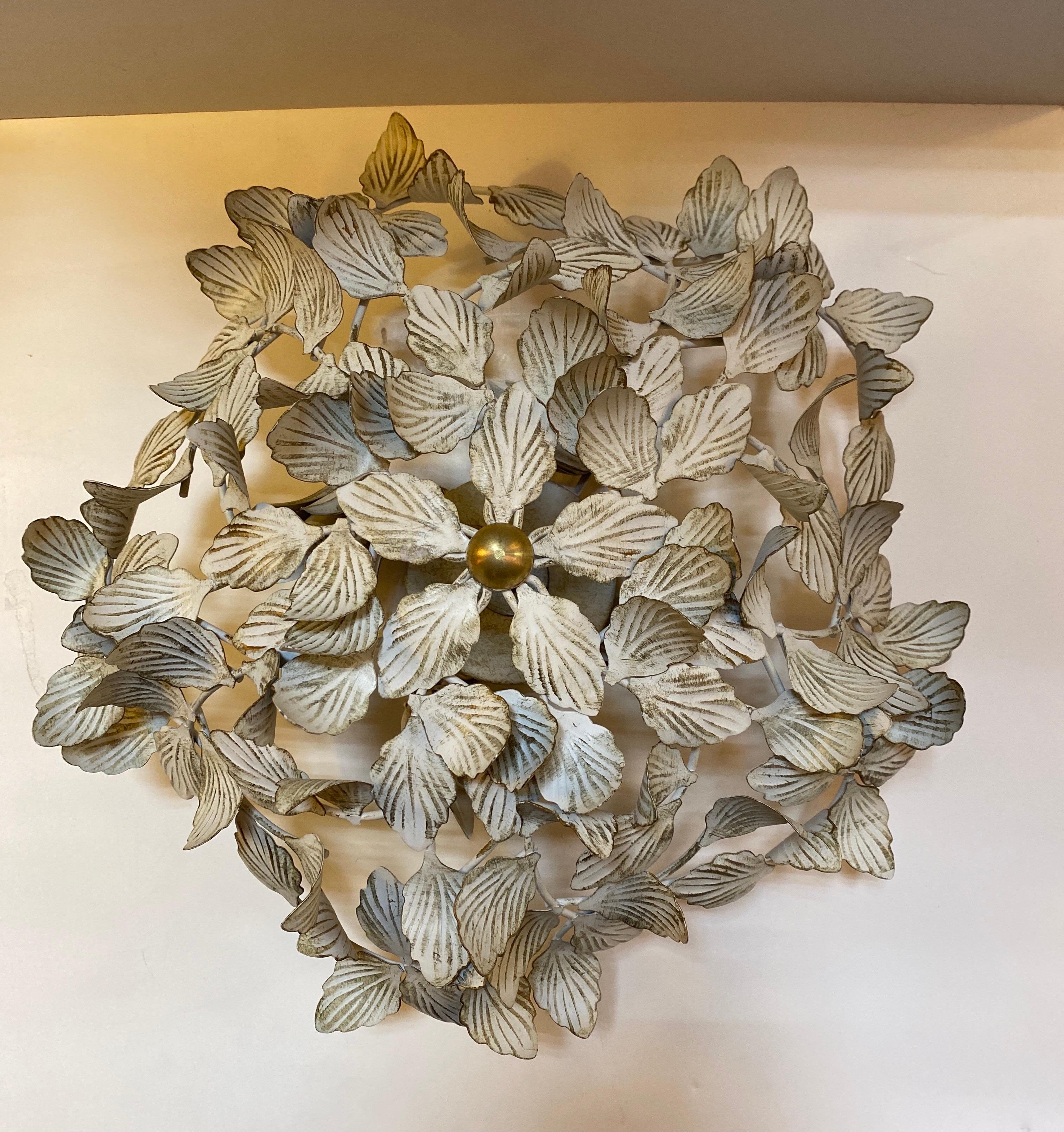 A vintage 4-light flush mount light fixture in the Hollywood Regency style. 

Features a round shape with fanciful ivy leaves in white tole, complemented by a brass finial.

USA, circa 1960.

Takes 4 (four) US candelabra base