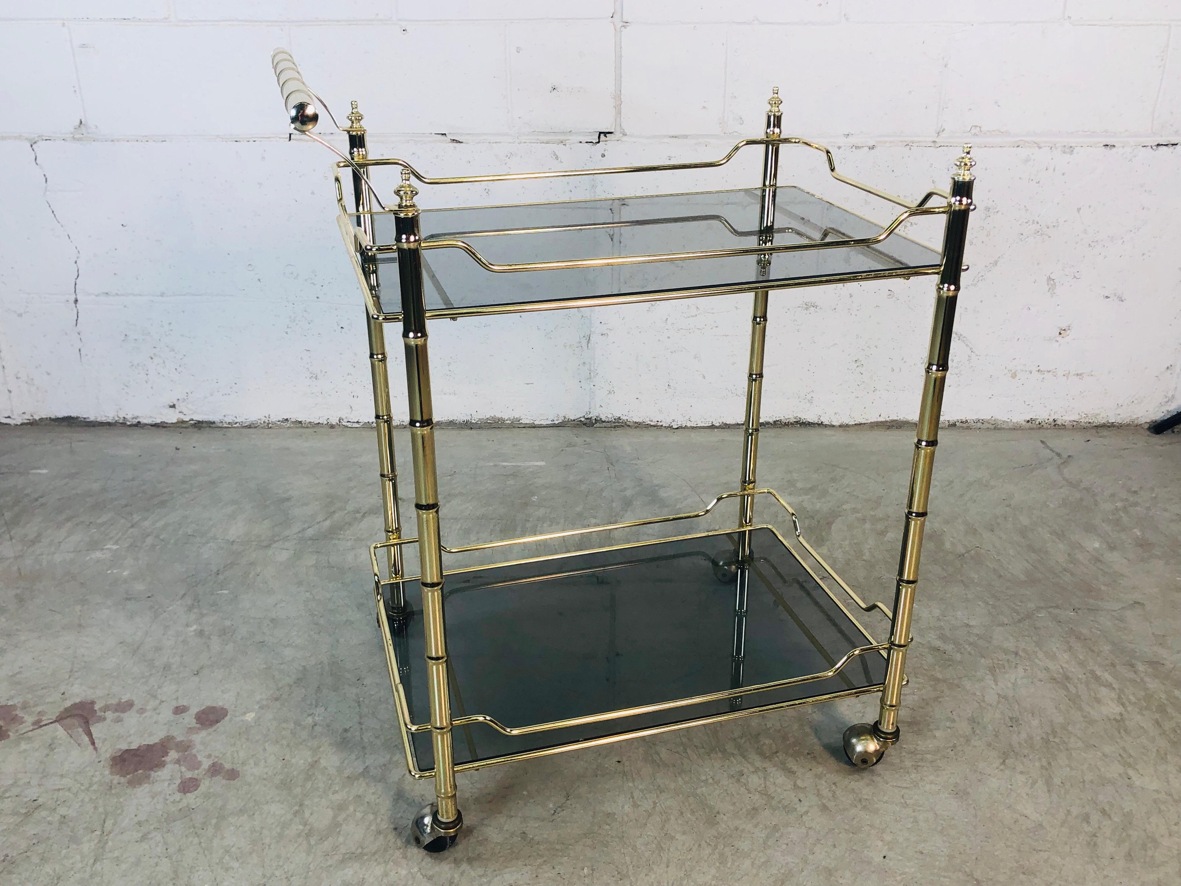 1970s Hollywood Regency gilt bamboo style rolling bar cart with smoked glass shelves. The cart is metal with glass shelves and rolls freely. Very good used condition. No makers mark.