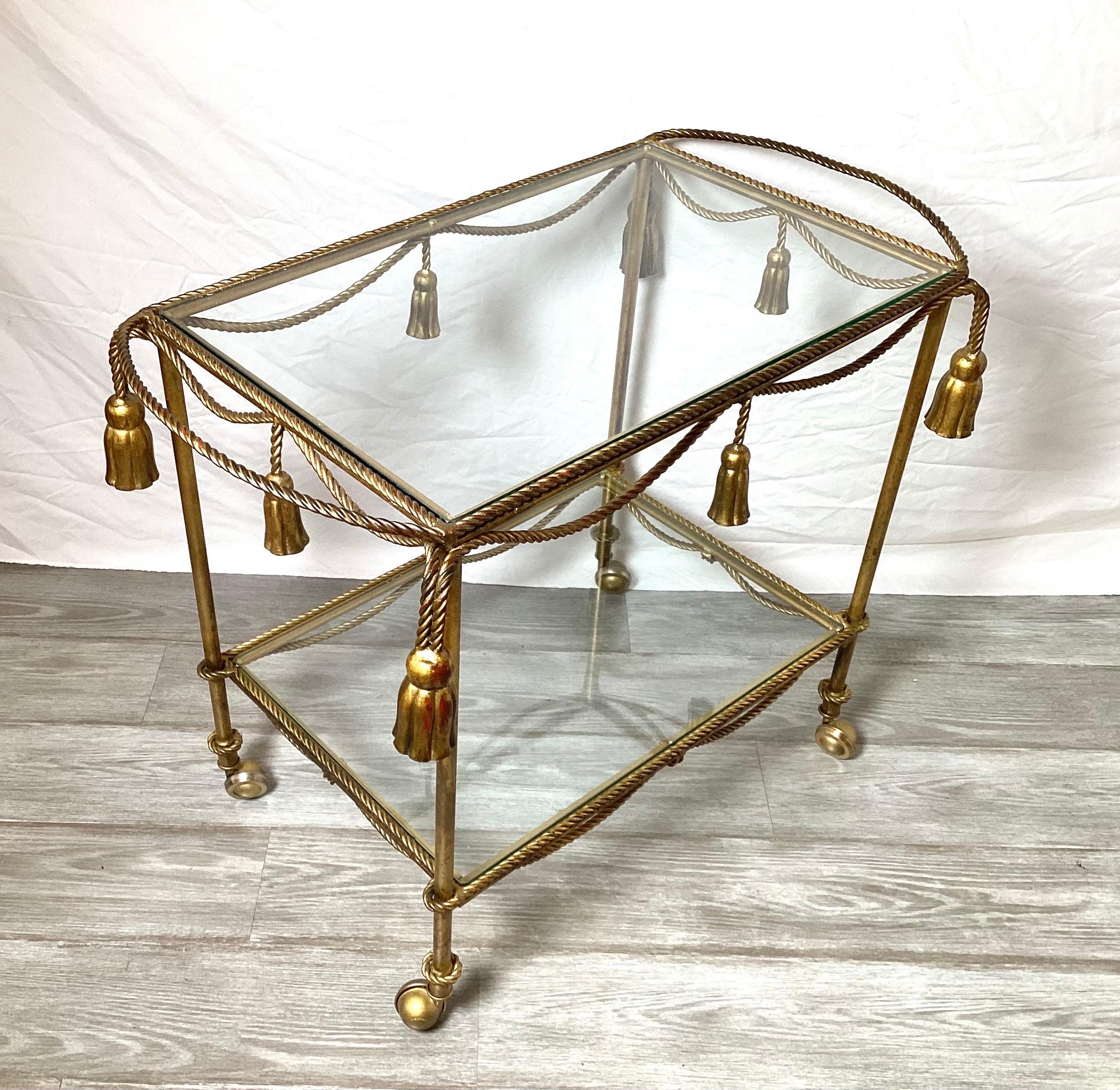A charming gilt metal and glass tiered bar dessert or beverage cart. The rope style trim with tassels with two shelves resting of castors.