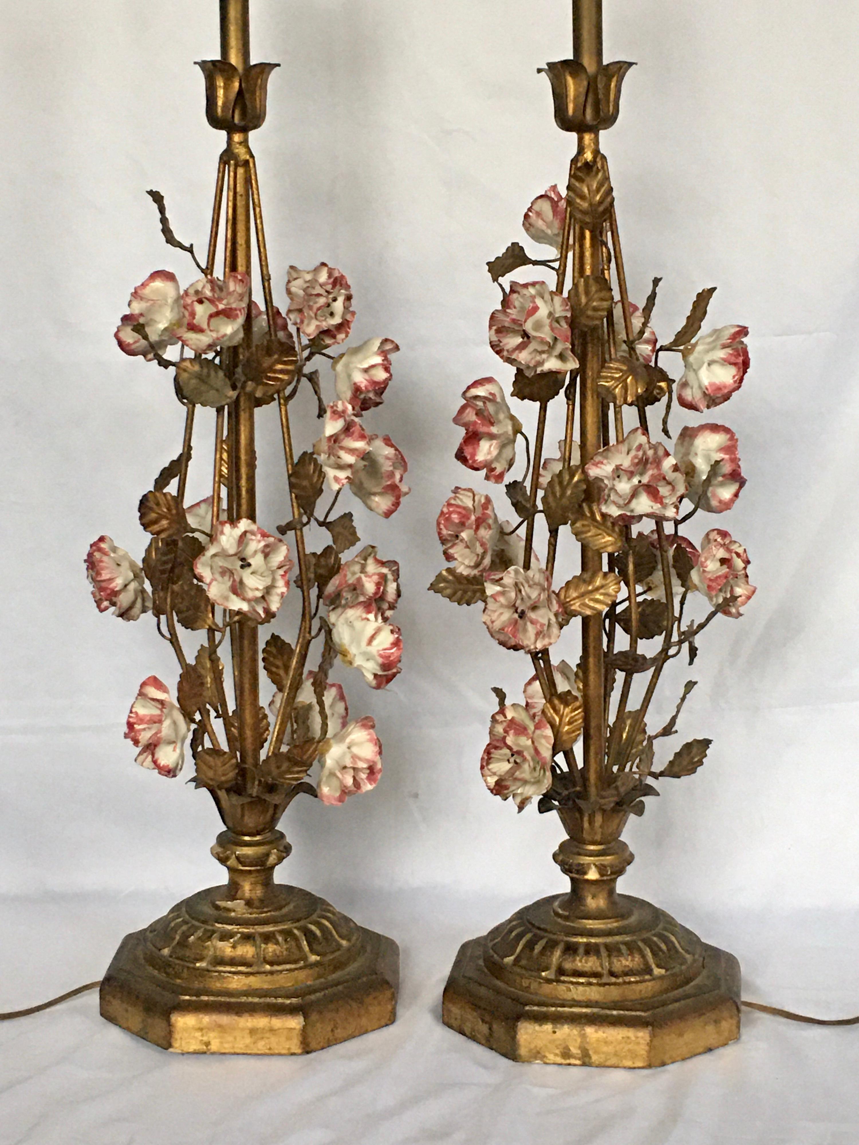 Pair of Hollywood Regency tole floral table lamps mounted on carved giltwood bases. These Italian lamps feature hand painted porcelain blooms with metal gilt vines and leaves. Bases marked 'Made In Italy'. Lamp shades not included. 

Height to