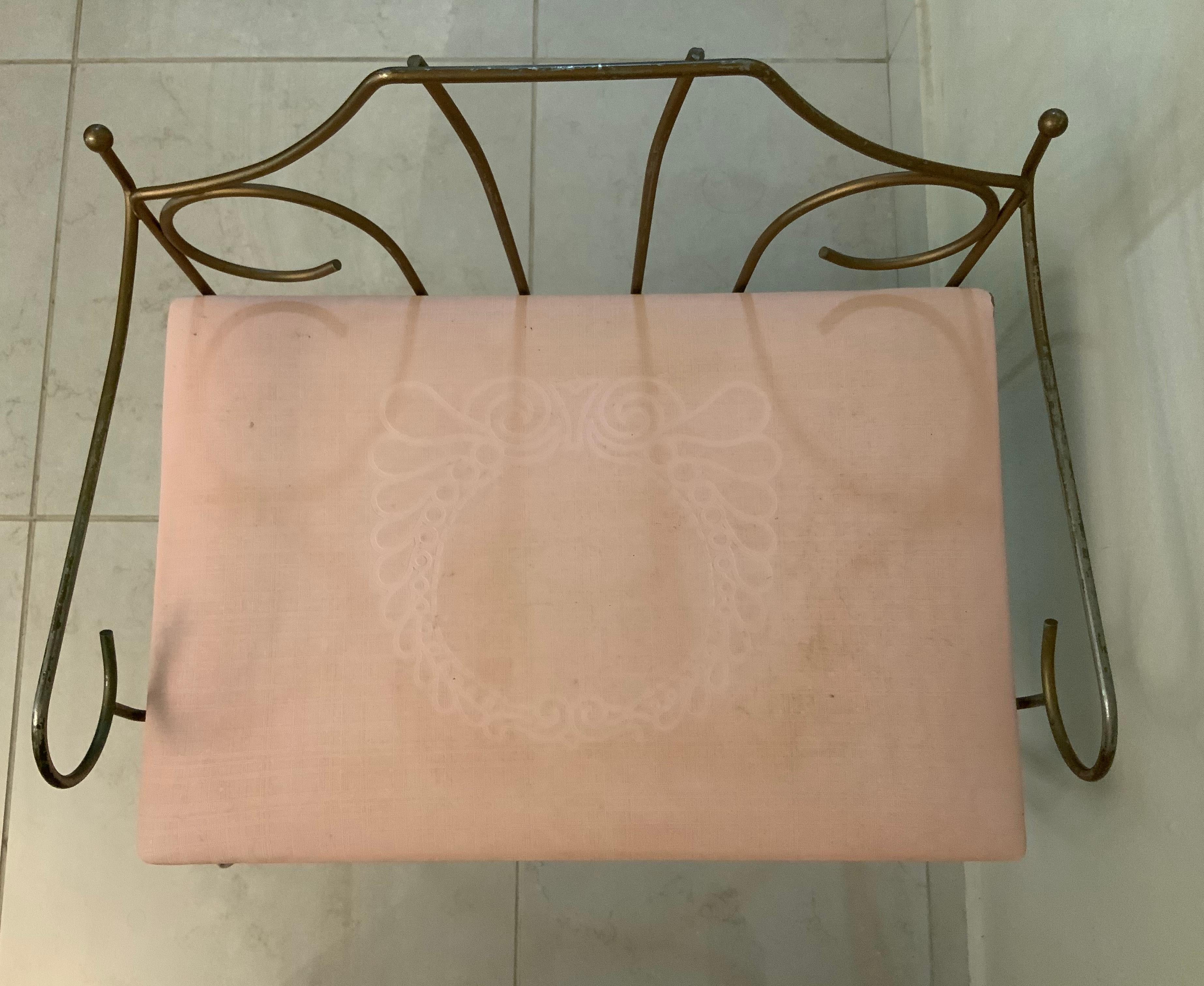 This is a gold tone metal vanity rectangular bench/stool. It has a pale pink vinyl seat that is adorned with a wreath. It has a pentagonal pierced metal back adorned with scrolls. It has also a scroll shaped metal arms. The bench is supported by