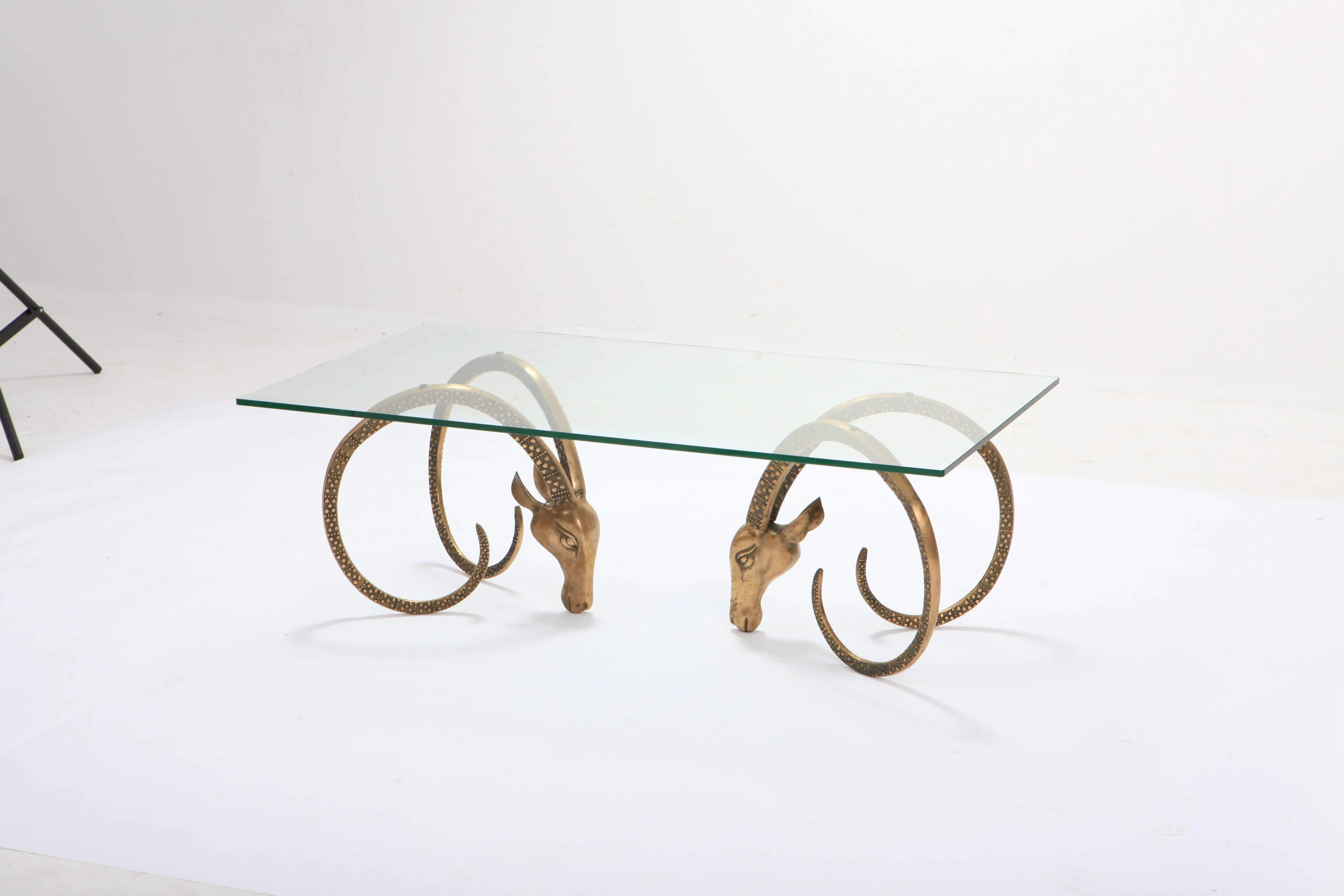 Brass Mid-Century Modern Ibex coffee table with a clear glass top.

Hollywood Regency style sculptural ram head bases with long patterned horns.

France, 1970s.
    