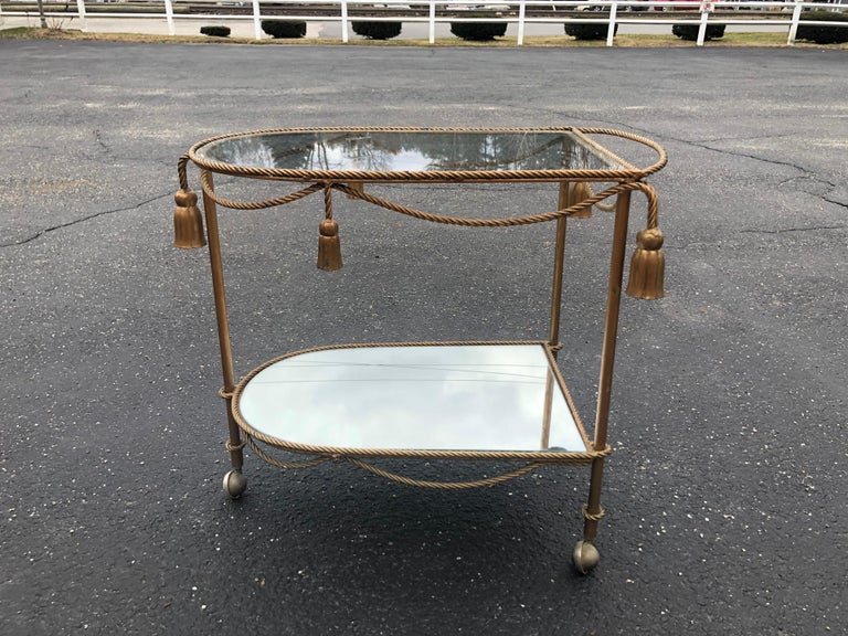 Hollywood Regency Italian bar cart. Stunning gilt iron piece with four tassles and intricate detailed decorative roping . Sexy and sleek. Mirrored bottom and clear glass top shelf. Interesting three wheel design. Perfect for entertaining.