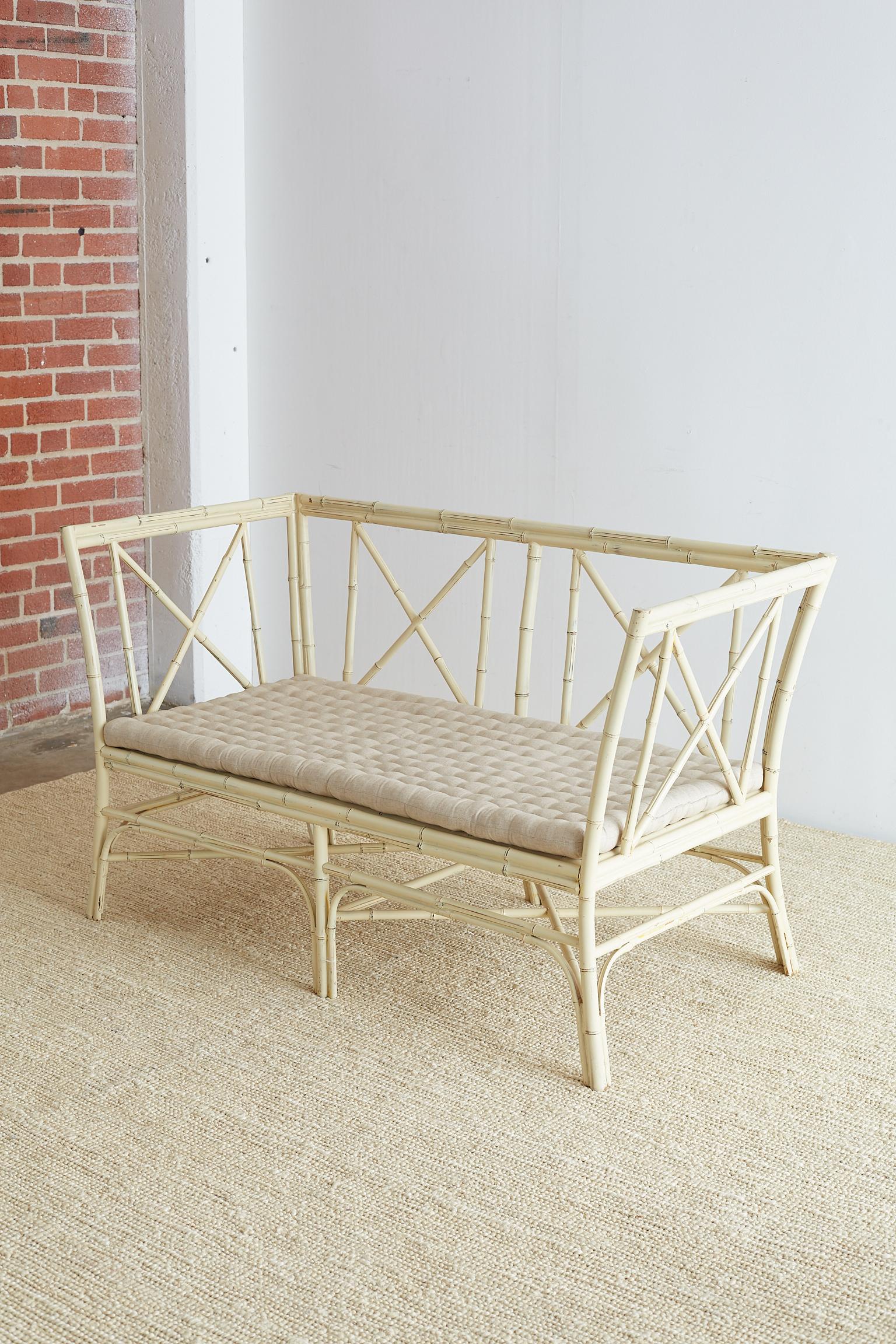 Elegant Hollywood Regency bamboo settee or bench featuring a cream colored lacquer finish. Delicate frame constructed from reeds of bamboo in a geometric pattern with X-form motif back, sides, and stretchers. Supported by six legs with a tufted seat