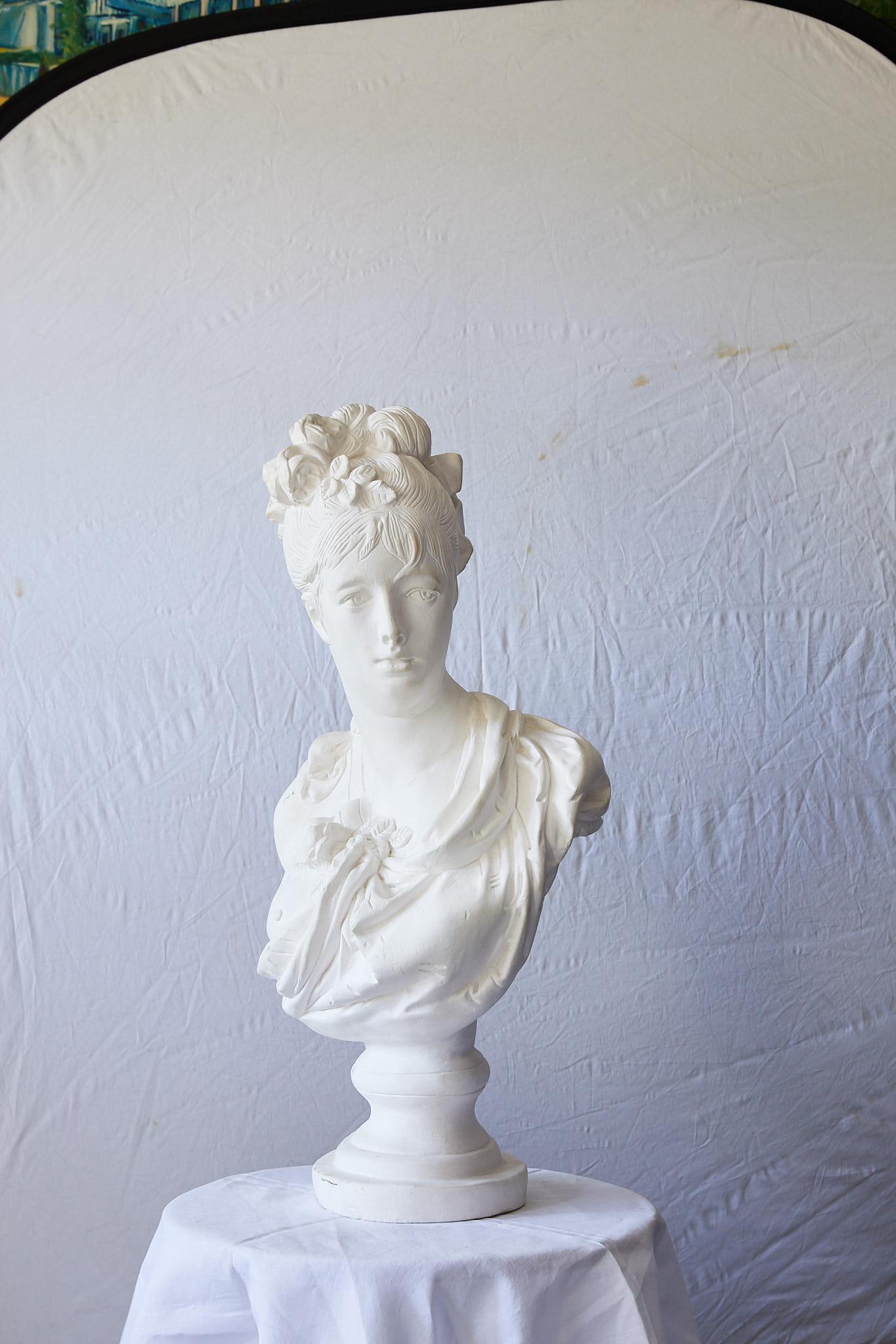 20th century large Hollywood Regency cast plaster portrait of a lady. Flowers and ribbons beautifully accent the upswept hair and draped fabric bodice. The vintage sculptural bust is raised on a tiered round pedestal and is signed on the back.