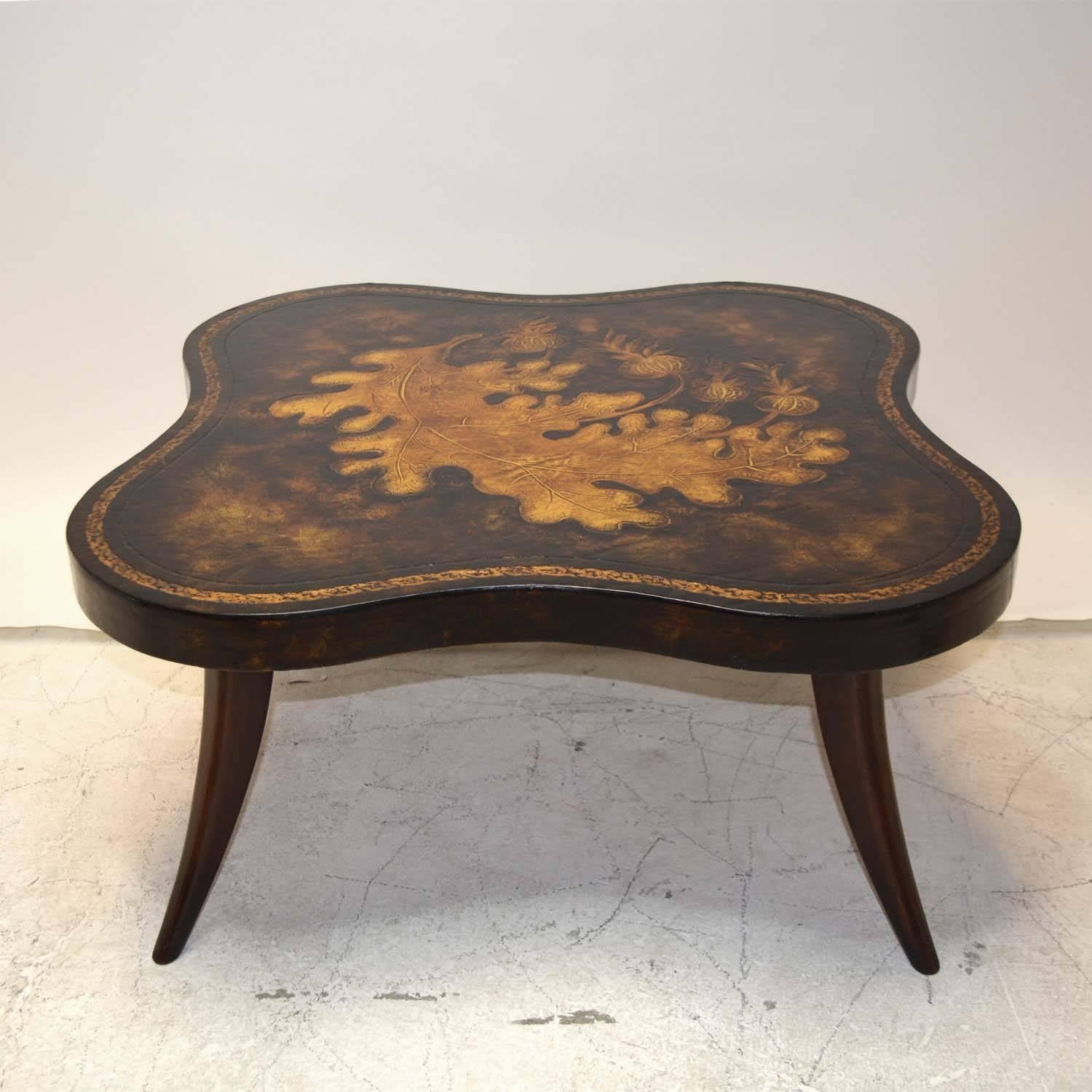 A Mid-Century Modern coffee table with incised leather top showing leaves and pomegranates, with gilt accented border, on saber legs. In the manner of Billy Haines and Paul Frankl.