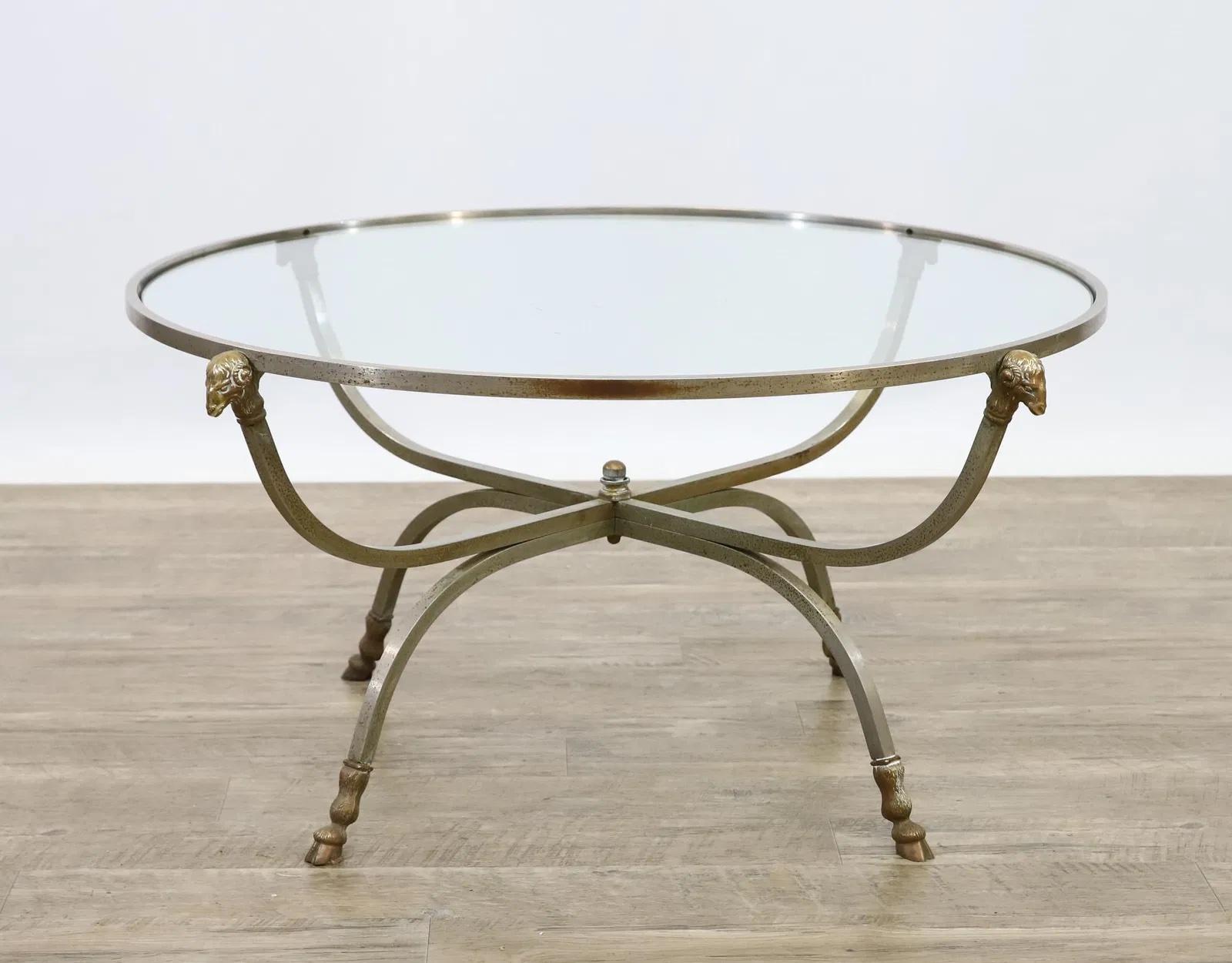 Hollywood Regency round coffee table with a steel and brass base featuring curved legs and supports decorated with ram monopodia, and an inset glass top.
