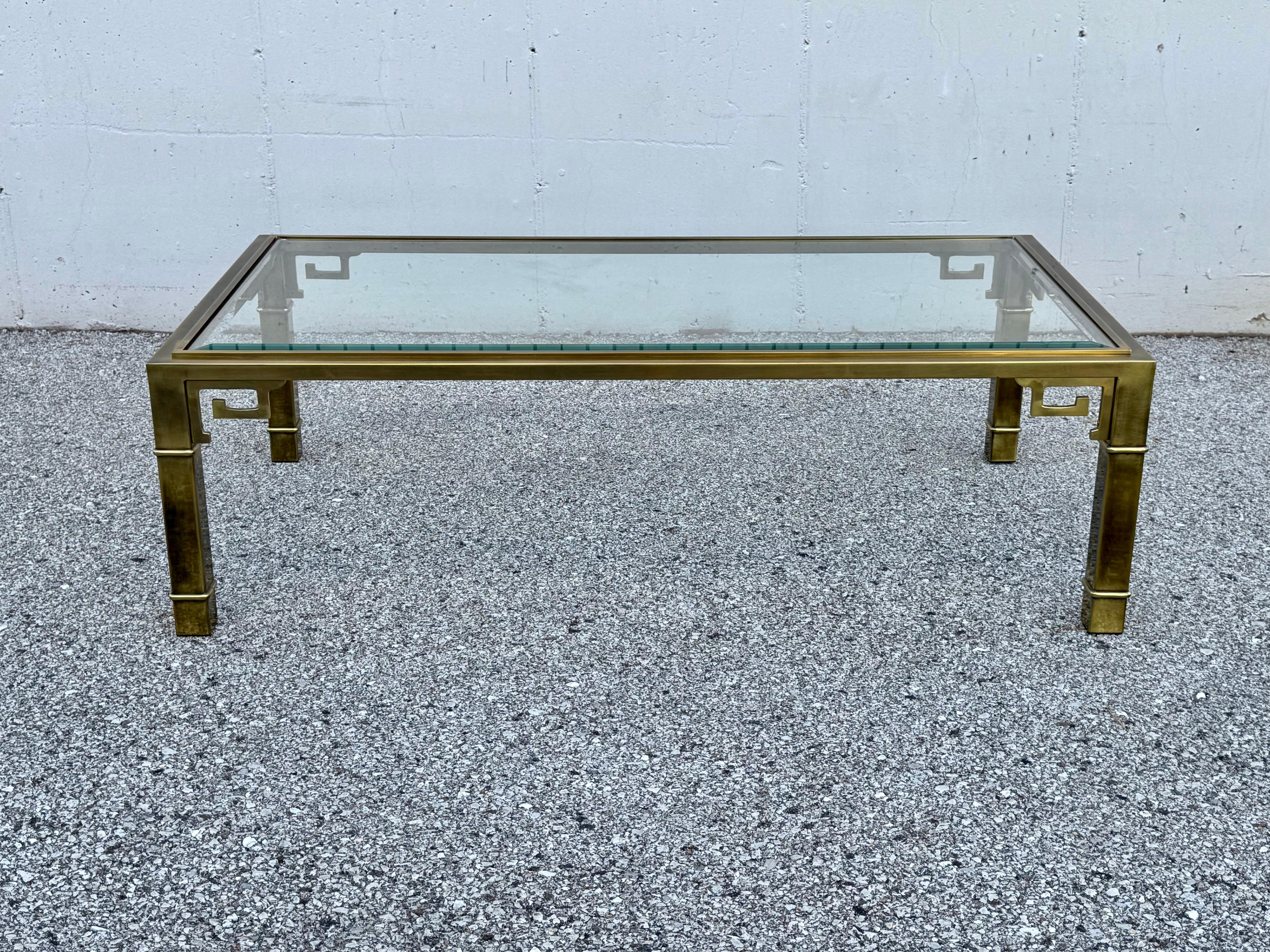 Table basse Greek Key de Mastercraft datant des années 1970, avec plateau en verre biseauté et cadre en laiton.  La base en laiton a une patine riche et chaude, le verre a quelques rayures et éclats dans les coins. Issue d'un domaine à propriétaire
