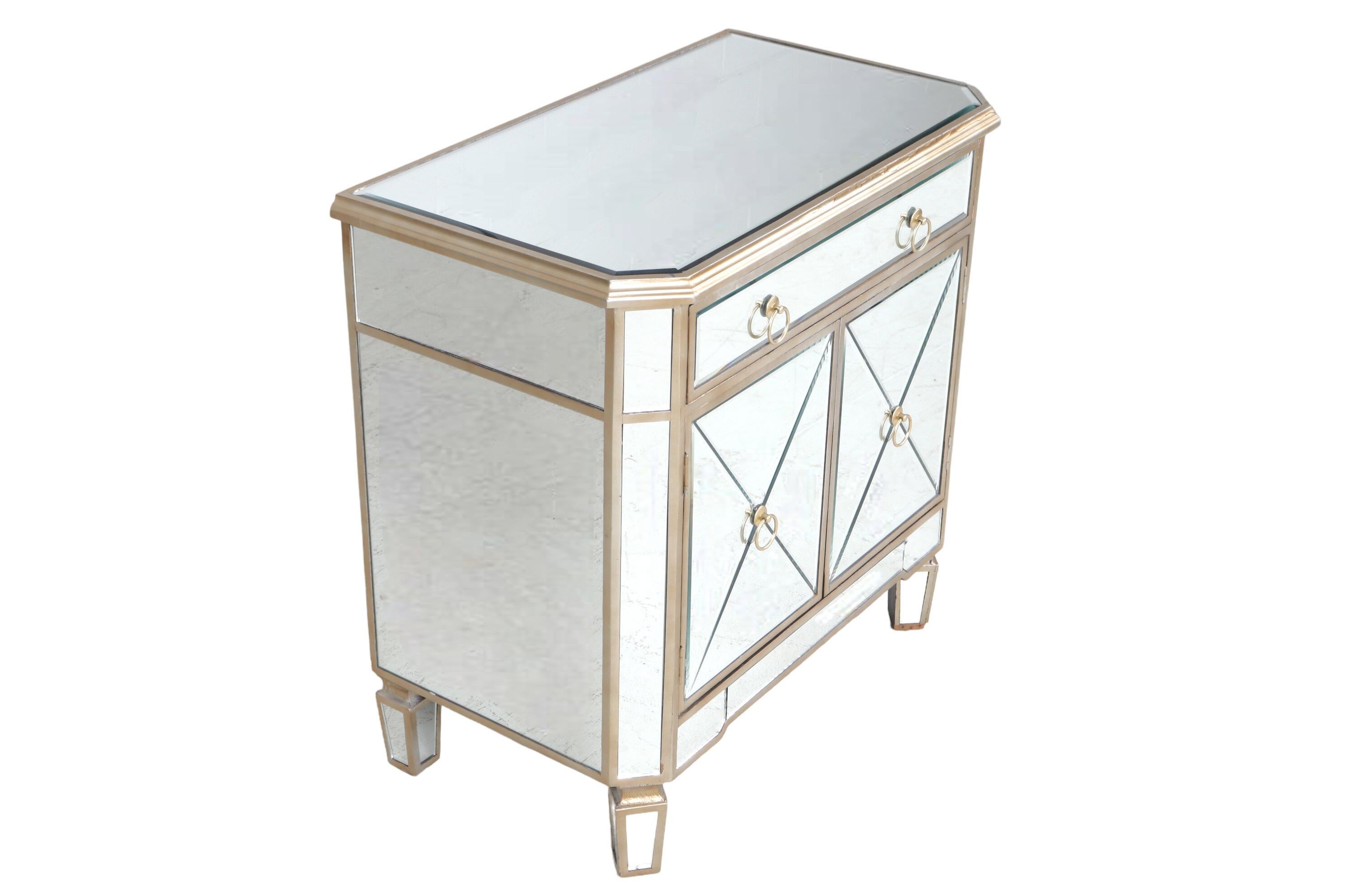 A Hollywood Regency style mirrored cabinet. Beveled mirrored glass panels cover each surface, framed with wood in a champagne finish. A single drawer and two cabinet doors below open with round loop handles. Stands on mirrored square tapered legs.