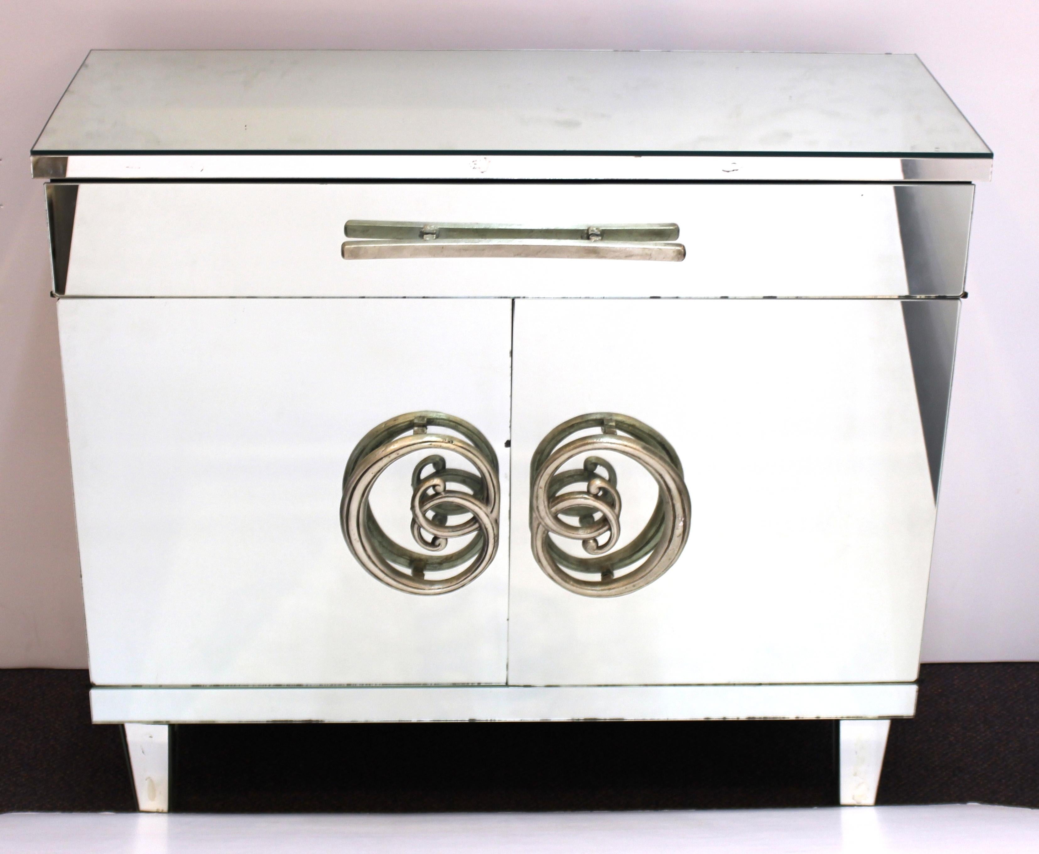 American Hollywood Regency pair of commodes with mirrored surfaces and ornate metalwork handles. The pair has each one large drawer atop a pair of hinged doors and ebonized wooden interior surfaces. In good vintage condition with some age-related