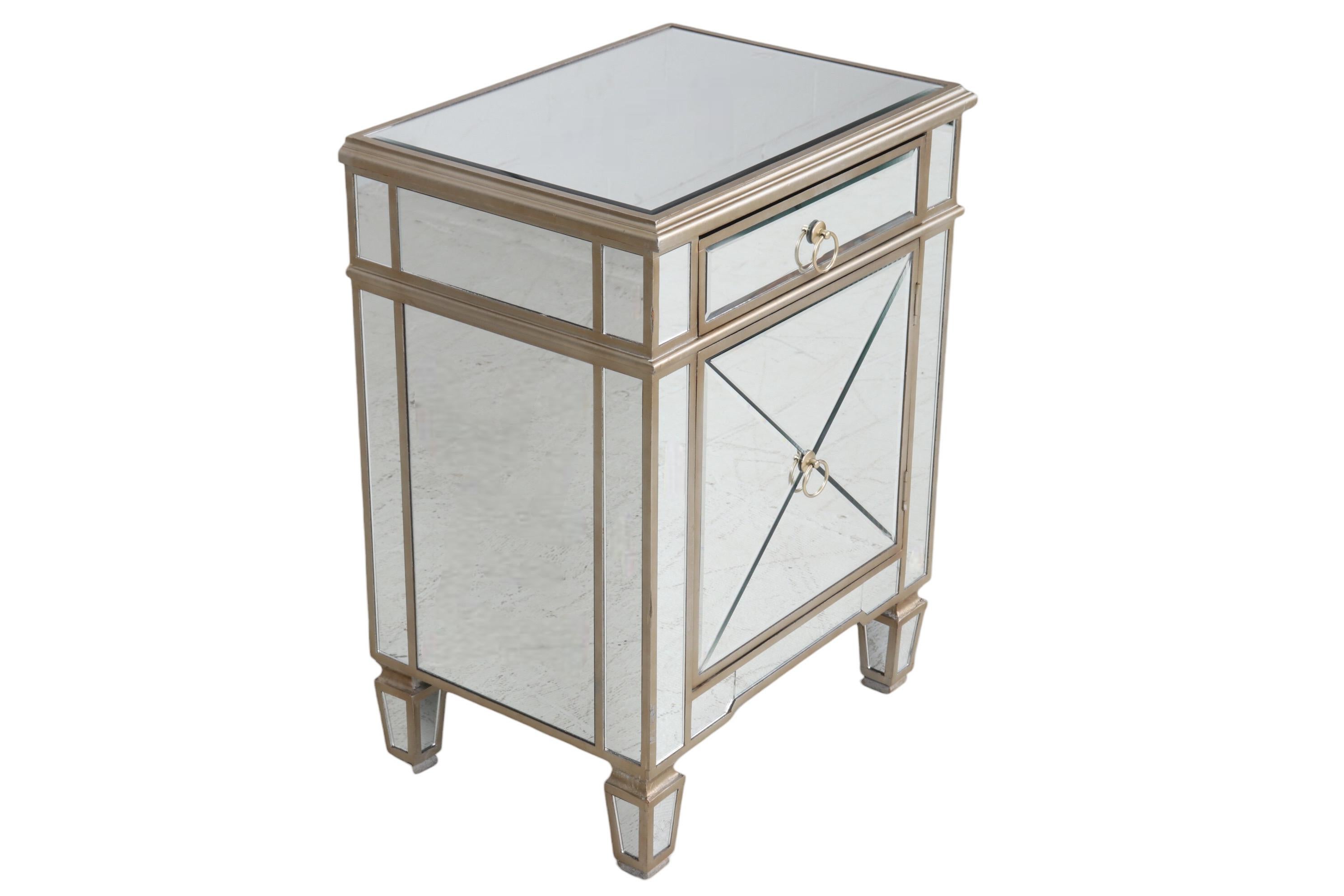 A Hollywood Regency mirrored cabinet, could be used as a nightstand or side table. Beveled mirrored glass panels cover each surface, framed with wood in a champagne finish. A single drawer and cabinet below open with round loop handles. Stands on
