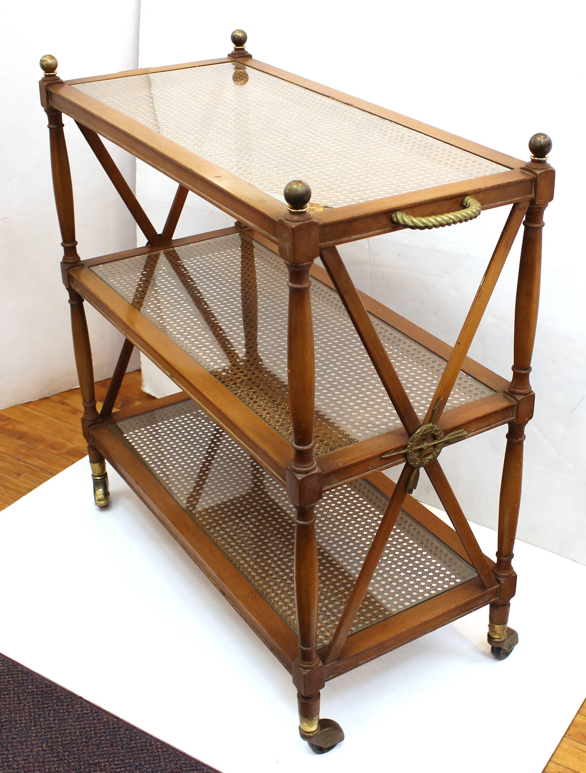 Bar cart produced during the 1940s in the neoclassical style. The cart features three tiers in cane, wood and glass. The piece also includes castors and brass details such as twisted rope handles and wreath medallions with arrows. Wear appropriate