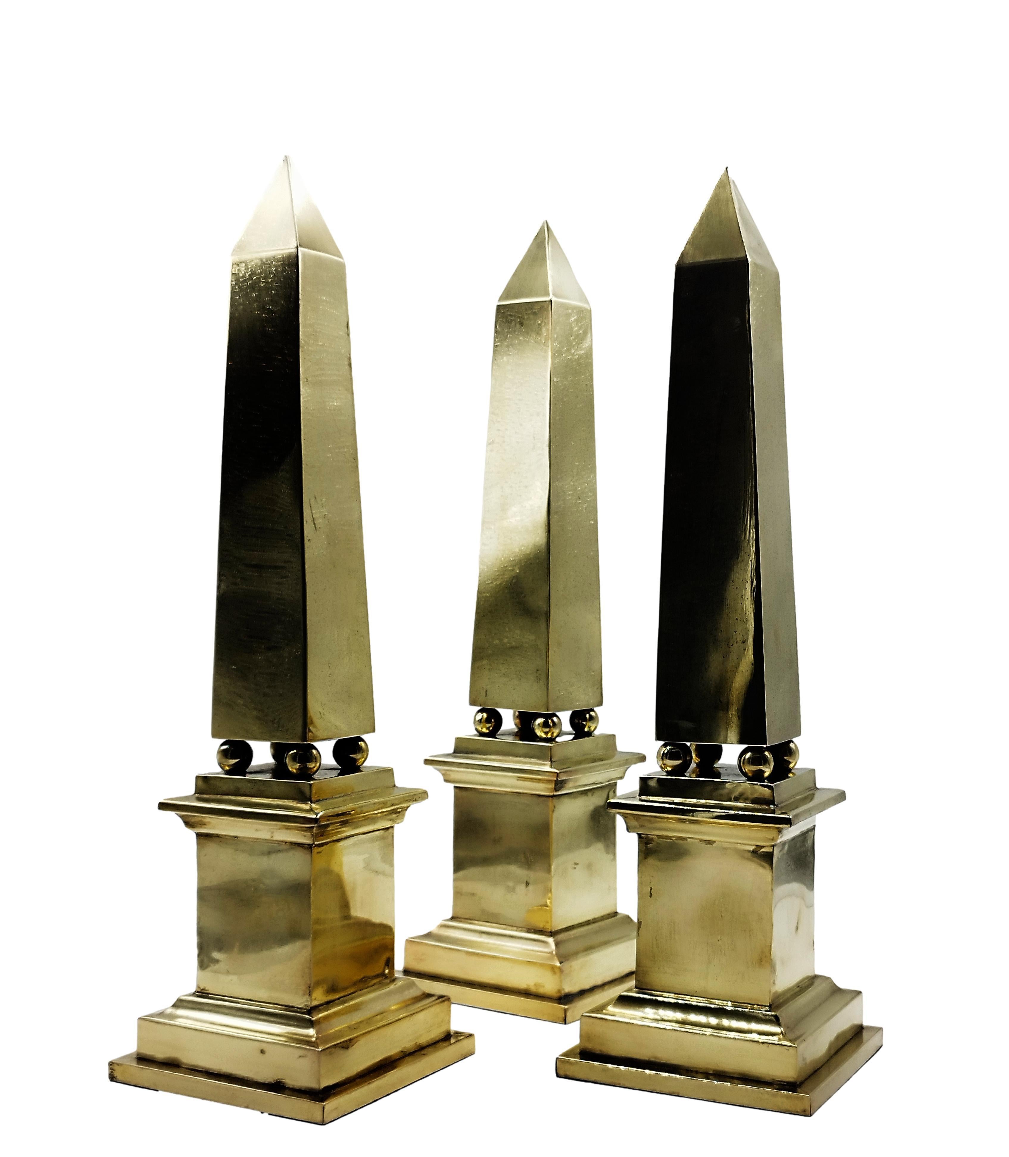 Brass obelisk with a square skyscraper-style base on which four spherical brass forms support the body. Obelisks are tapering rectangular volumetric shapes with faceted tops that come to a slender point. Harking back to classical Greek forms and