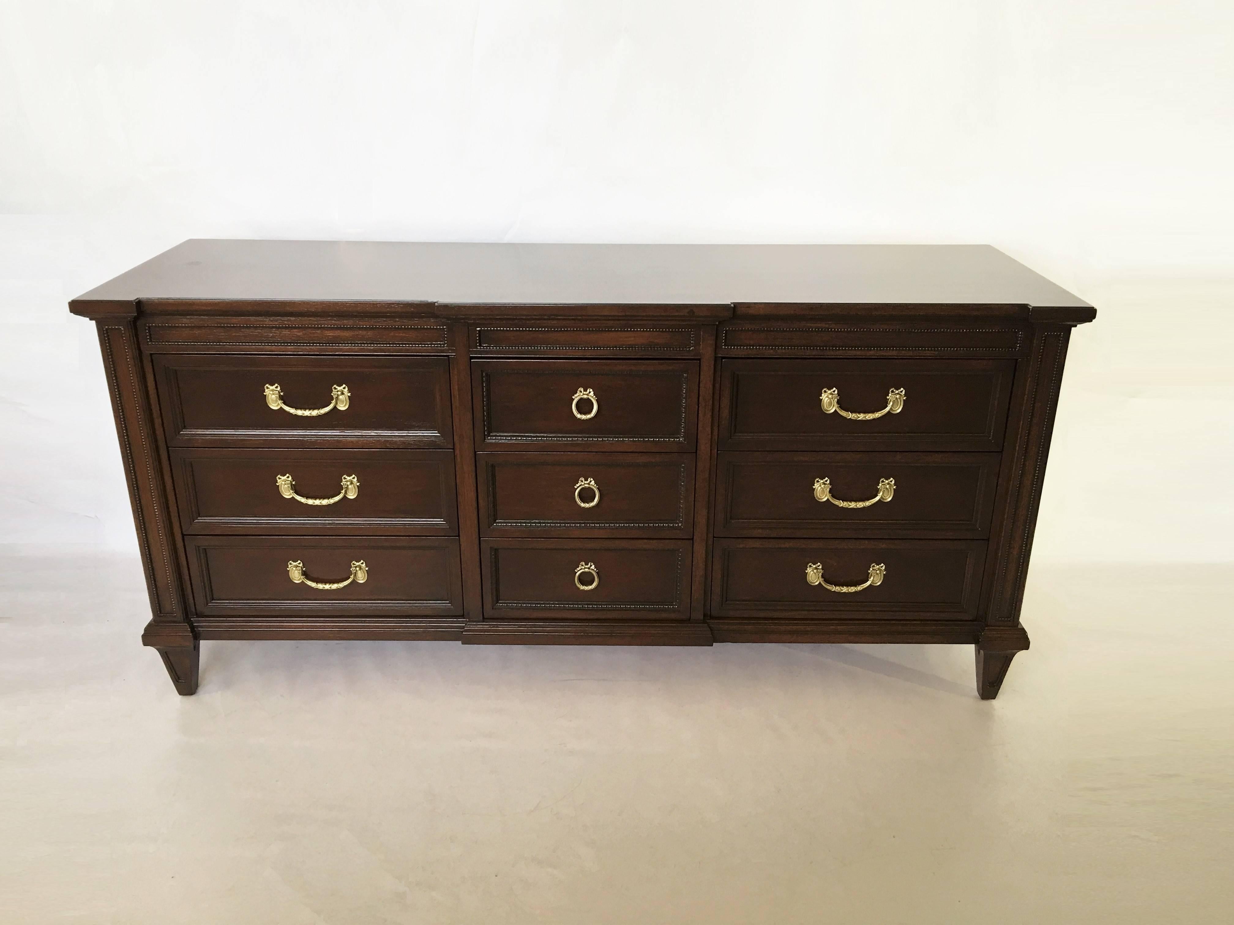 This 1970s vintage Regency-style dresser is made of mahogany wood that has been finished in a rich dark stain. The facade of the chest features nine drawers in three rows with ample storage space on tapered legs. The drawers have decorative brass